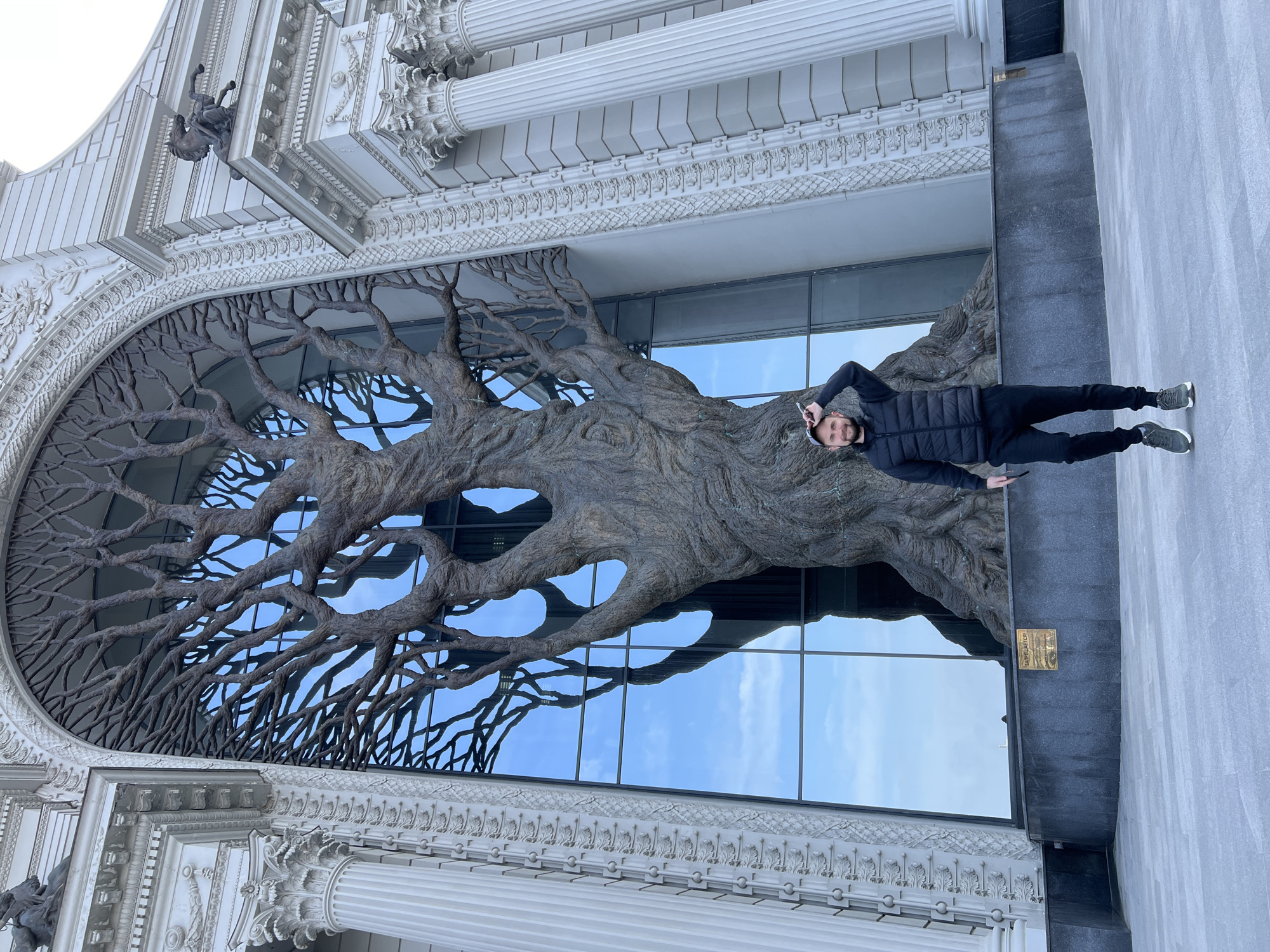 A medium-angle, exterior shot depicts a man standing in front of the Farmer's Palace in Kazan, Russia. The building facade features an arched, ornate window with a large, sculptural tree embedded in it. The man, dressed in a black puffer jacket and black pants with gray sneakers, faces the camera with his head tilted and his left arm raised, holding what appears to be a cell phone to his ear. The building is constructed in a grand, palatial style with white architectural details, columns, and sculptural embellishments. The sky is visible through the glass behind the tree.
