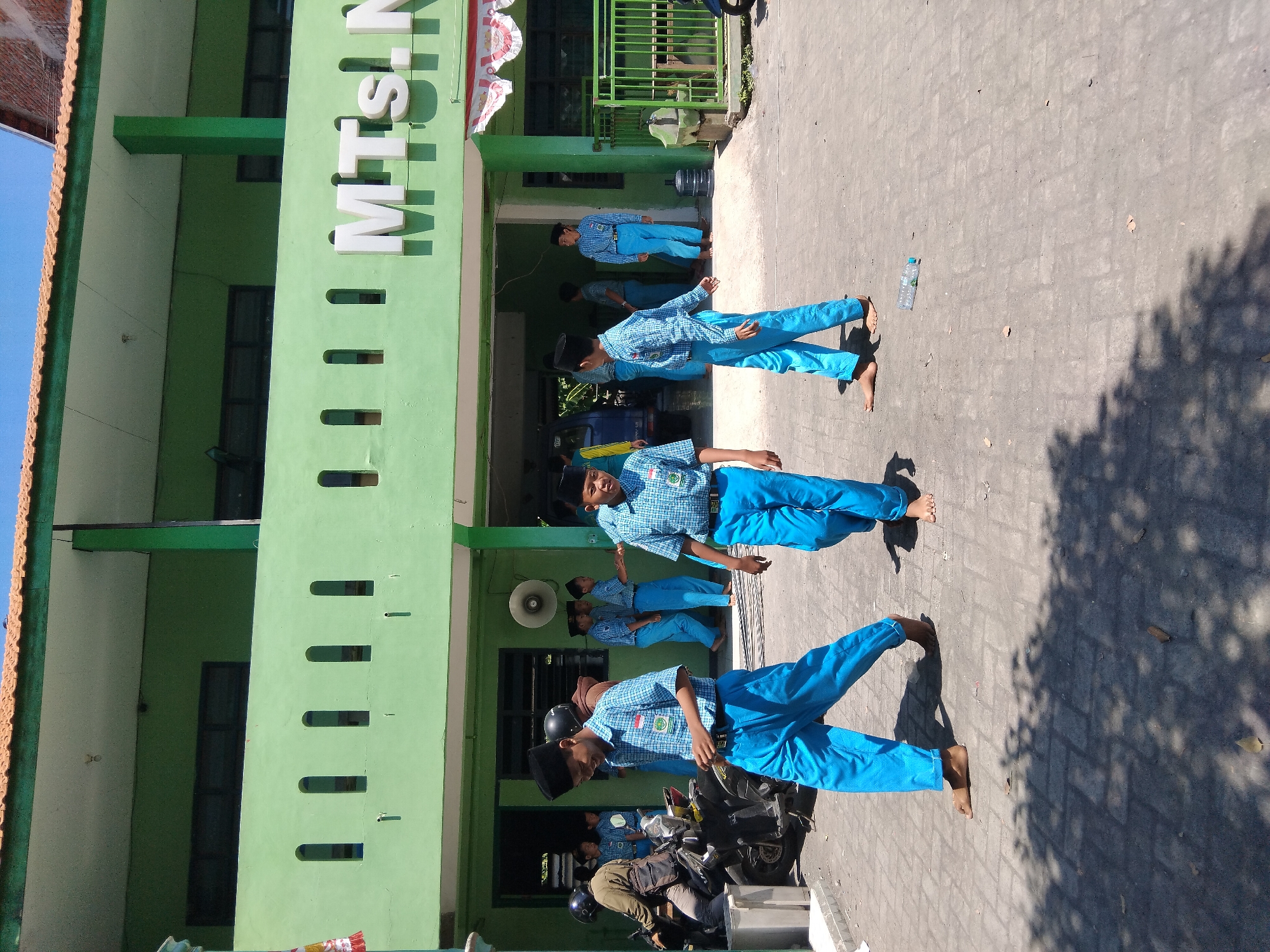 In Kepuh Kiriman, Indonesia, a group of young boys in blue and white checkered uniforms and black peci hats are present outside a light green, two-story building with a tiled roof. The building has the white lettering "MTs. N" visible on its facade. Some of the boys are walking on a paved area, while others are standing. In the foreground, two boys are walking towards the camera, one smiling and looking to the right. To the left, a person in a dark jacket and helmet is seated on a motorcycle. The environment appears to be sunny, with shadows cast by trees and the building onto the paved ground. A plastic water bottle is visible on the ground in the middle ground.