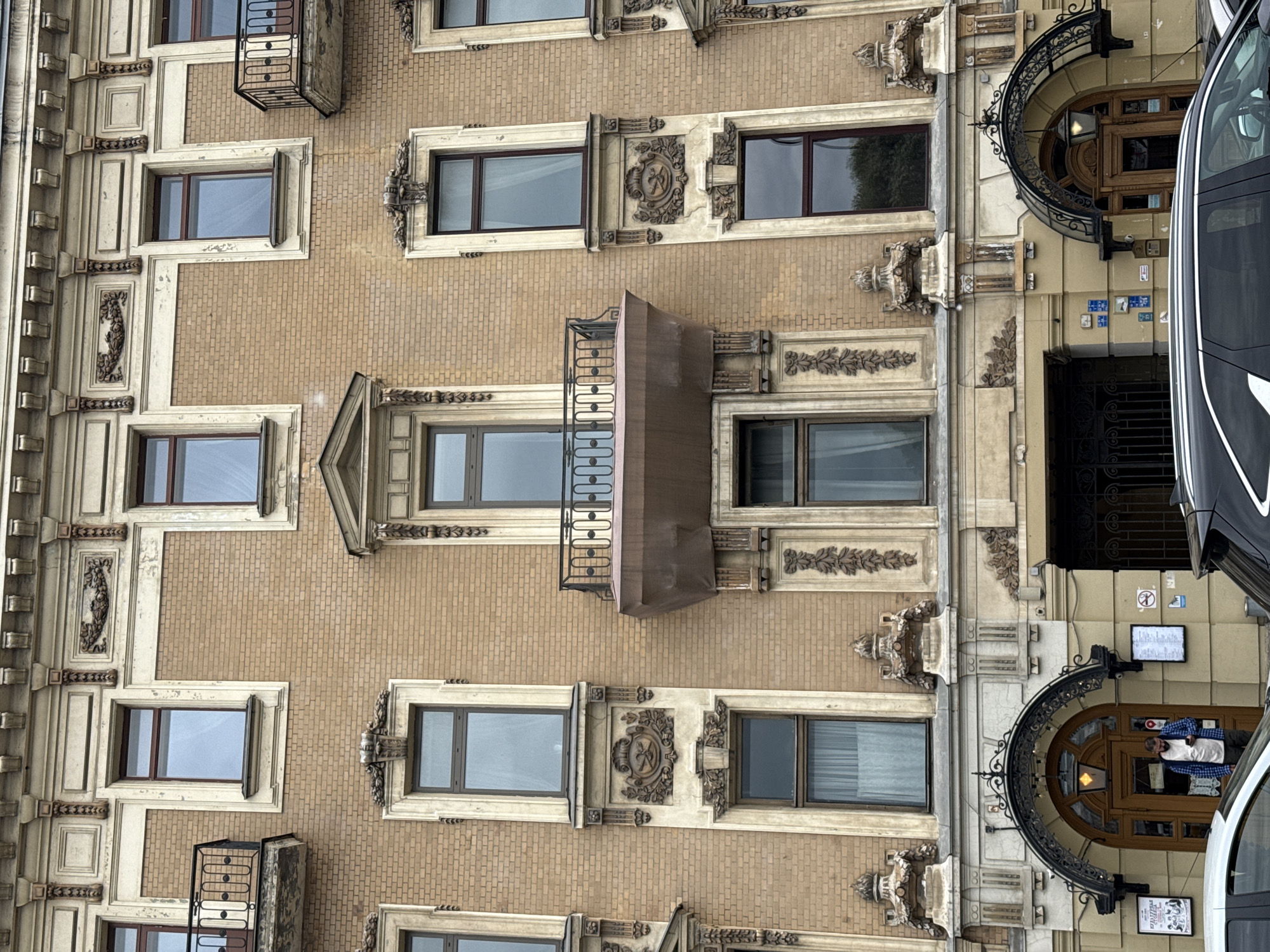 A front-facing, eye-level exterior medium shot captures a section of a multi-story building with ornate architectural details. The building features a light brown brick facade with cream-colored decorative elements around the windows and along the roofline. Several windows, some with balconies and wrought-iron railings, are visible on the upper floors. The ground floor has a large arched entrance with a wooden door and a decorative metal awning. A man in a blue plaid shirt and white t-shirt stands in the doorway, looking at his phone. Two cars are parked in front of the building. The sky is overcast, suggesting a cloudy day. The overall impression is of an older, well-maintained urban building.