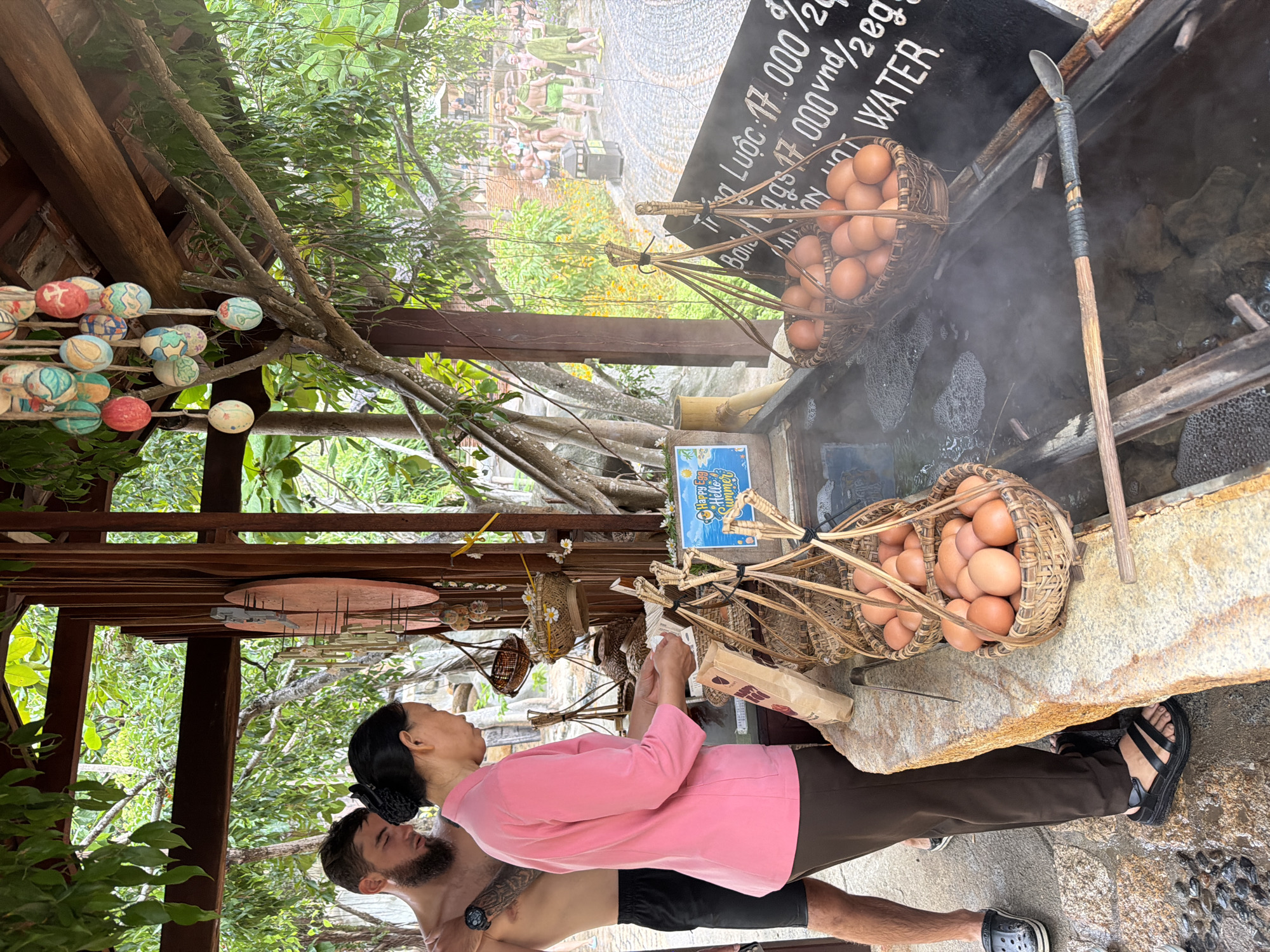 The image captures a scene at a hot spring egg boiling station in Nha Trang, Vietnam. In the foreground, a woman in a pink top and brown pants stands beside a stone counter, holding a small white object, possibly a ticket or receipt. A man with a beard, shirtless and wearing black shorts, stands to her left, looking down. Two woven baskets filled with brown eggs are suspended over a steaming hot spring. A blackboard behind the baskets displays pricing information in Vietnamese. In the background, people are seen relaxing in the hot springs, some wearing green wraps. The area is surrounded by lush greenery and appears to be an outdoor tourist attraction.