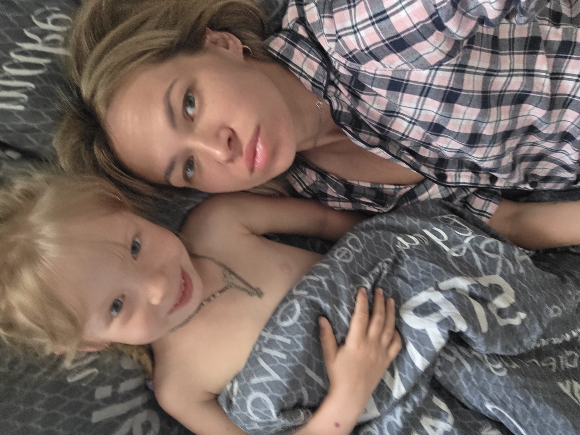 The image shows a fair-skinned woman and a fair-skinned child lying on a gray bed with a patterned gray and white blanket. The woman is wearing a pink, white, and blue plaid shirt. The child is partially covered by the blanket and wearing a necklace. The location is potentially Chaikovsky, Russia.