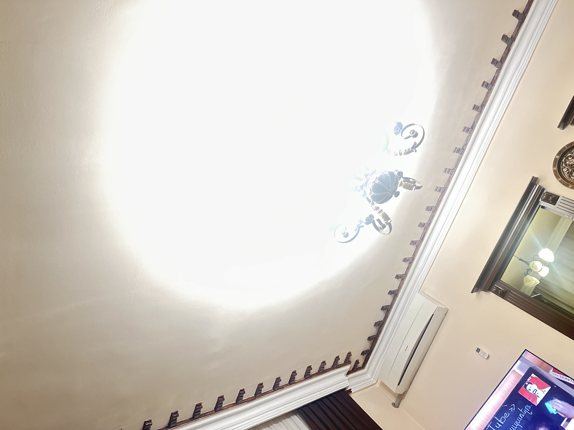 The image depicts an interior view looking up at a ceiling and adjacent wall. The ceiling is white with ornate brown trim around the edges. A chandelier hangs from the center of the ceiling. The wall is pale peach, with an air conditioning unit mounted on it. A framed mirror is visible, reflecting another light fixture. A television screen is visible in the corner of the image, displaying content. Part of a dark brown curtain is also visible. While I can't definitively confirm the location as Yerevan, Armenia based solely on the image, the presence of Armenian text on the television screen ("Tube >> հայկական" which translates to "Tube >> Armenian") suggests the image was taken in a location where Armenian is spoken. Without additional evidence, it's impossible to conclusively confirm Yerevan as the specific location.