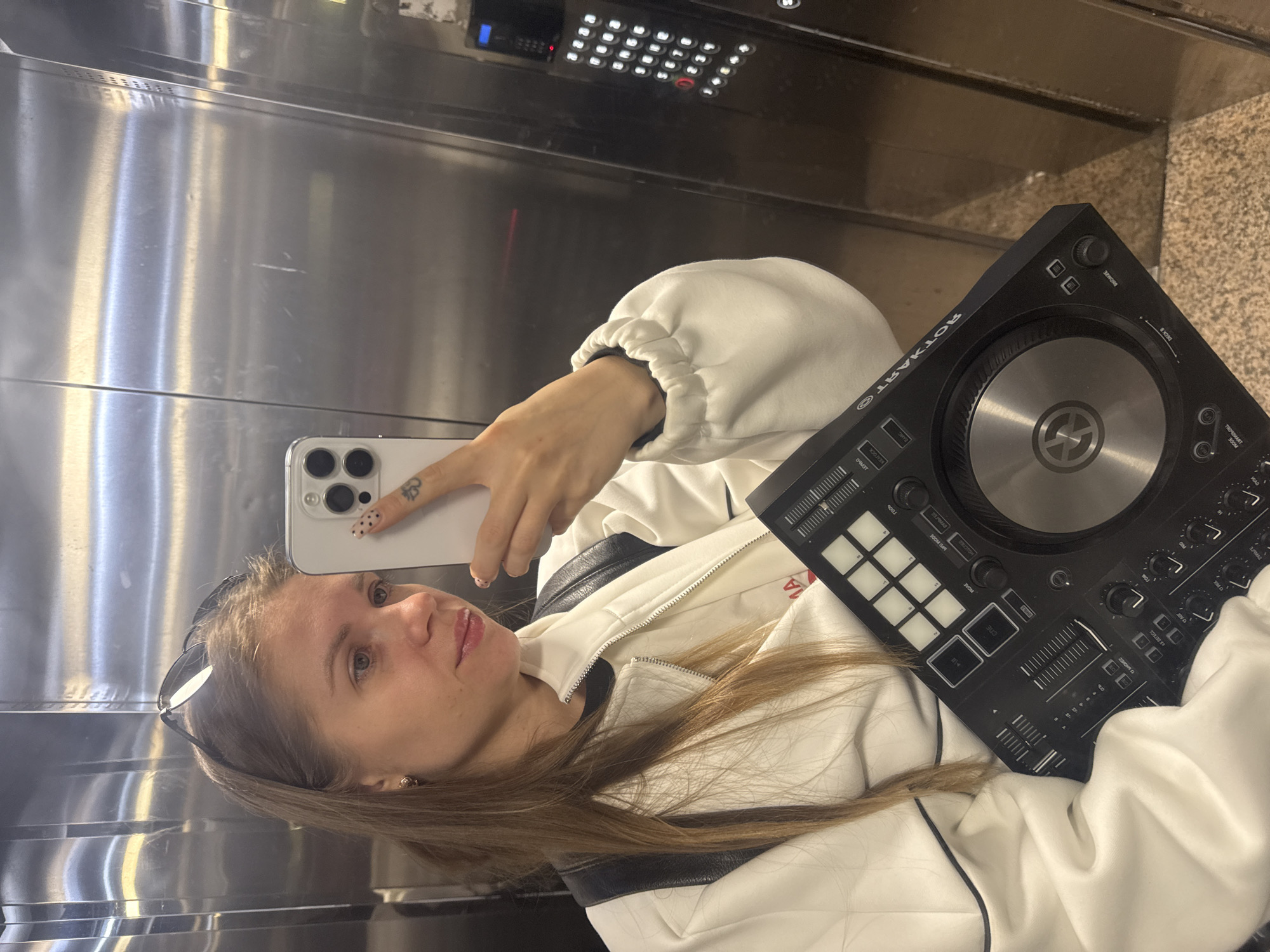 A woman is taking a selfie in an elevator. She is holding a white smartphone in her left hand, which is decorated with small, patterned stickers. Her right hand is holding a black Rotart electronic DJ mixer. She is wearing a white jacket with black trim. In the background, the elevator's brushed metal walls and control panel with numbered buttons are visible. Based on the available information, it is not possible to ascertain whether the location is in the city of Batumi, Georgia.