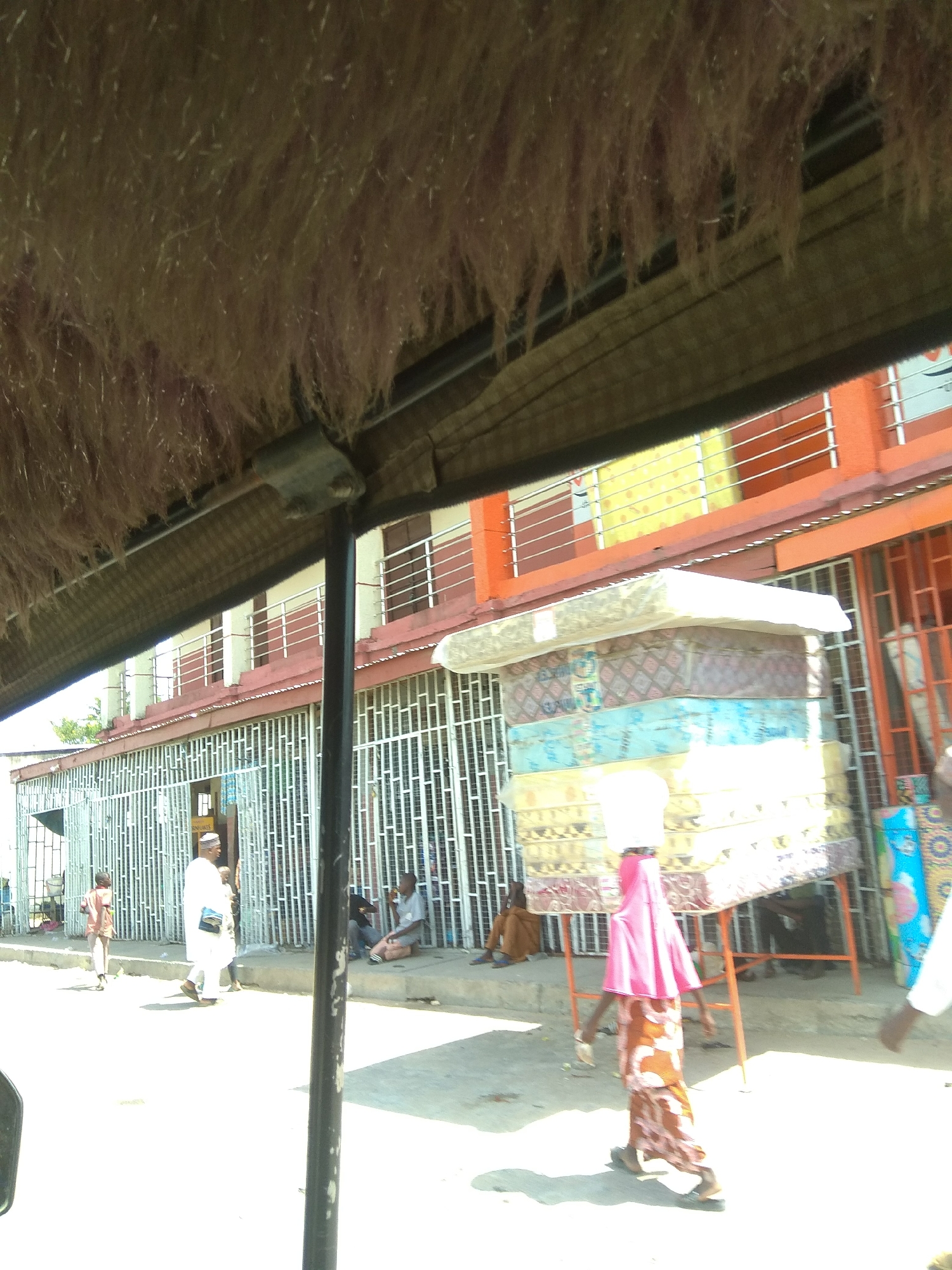 This image depicts a street scene in Jalingo, Nigeria. A woman in a pink headscarf and a patterned skirt is carrying a stack of mattresses on her head, walking towards the right. The mattresses are wrapped in plastic and are various colors and patterns. Behind her, a rack supporting the mattresses is visible. In the background, a building with orange and yellow accents and metal railings is present. A man in a white robe and cap is walking towards the left, and a child in an orange shirt is walking away from the camera. Several other individuals are seated or standing along the street, some behind metal bars that appear to be part of shopfronts or enclosures. The foreground is partially obscured by what appears to be a thatched roof or overhang with hanging fibers, possibly from a shelter or vehicle. The street is paved, and sunlight casts shadows on the ground.