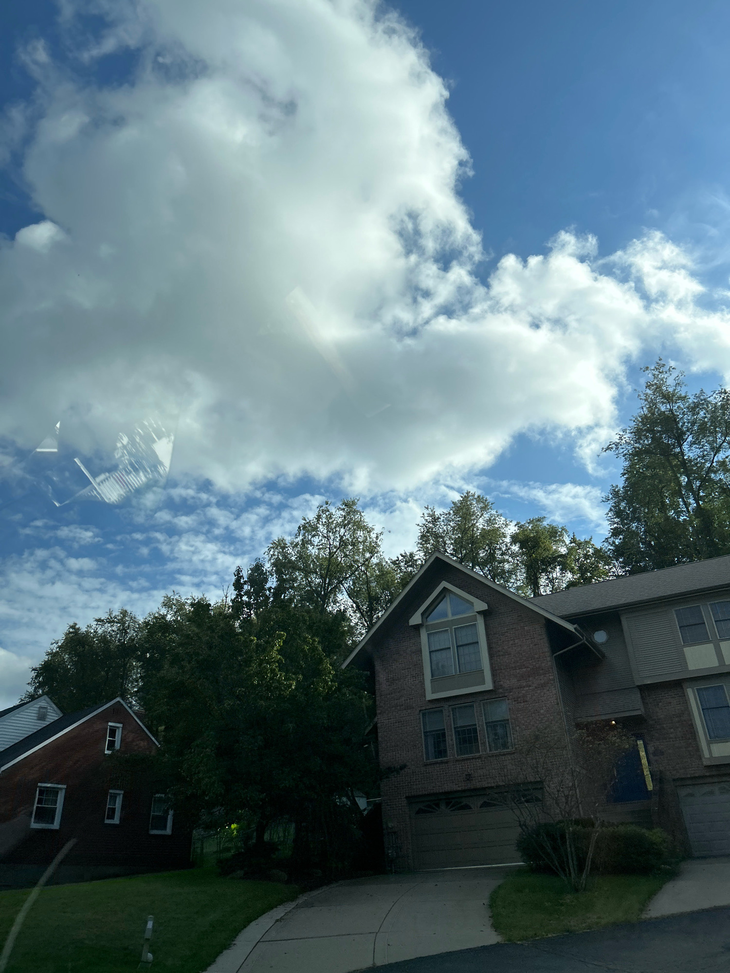 The image captures a residential street scene with two houses visible, nestled amongst lush green trees. The sky above is a vibrant blue, dotted with fluffy white clouds, indicating fair weather. In the foreground, a paved driveway leads up to a large brick house with a garage. To the left, a smaller brick house with white trim and windows is partially visible, set back from the street. The overall impression is one of a peaceful suburban neighborhood. The image does not provide specific details about the city of Pittsburgh, United States of America.
