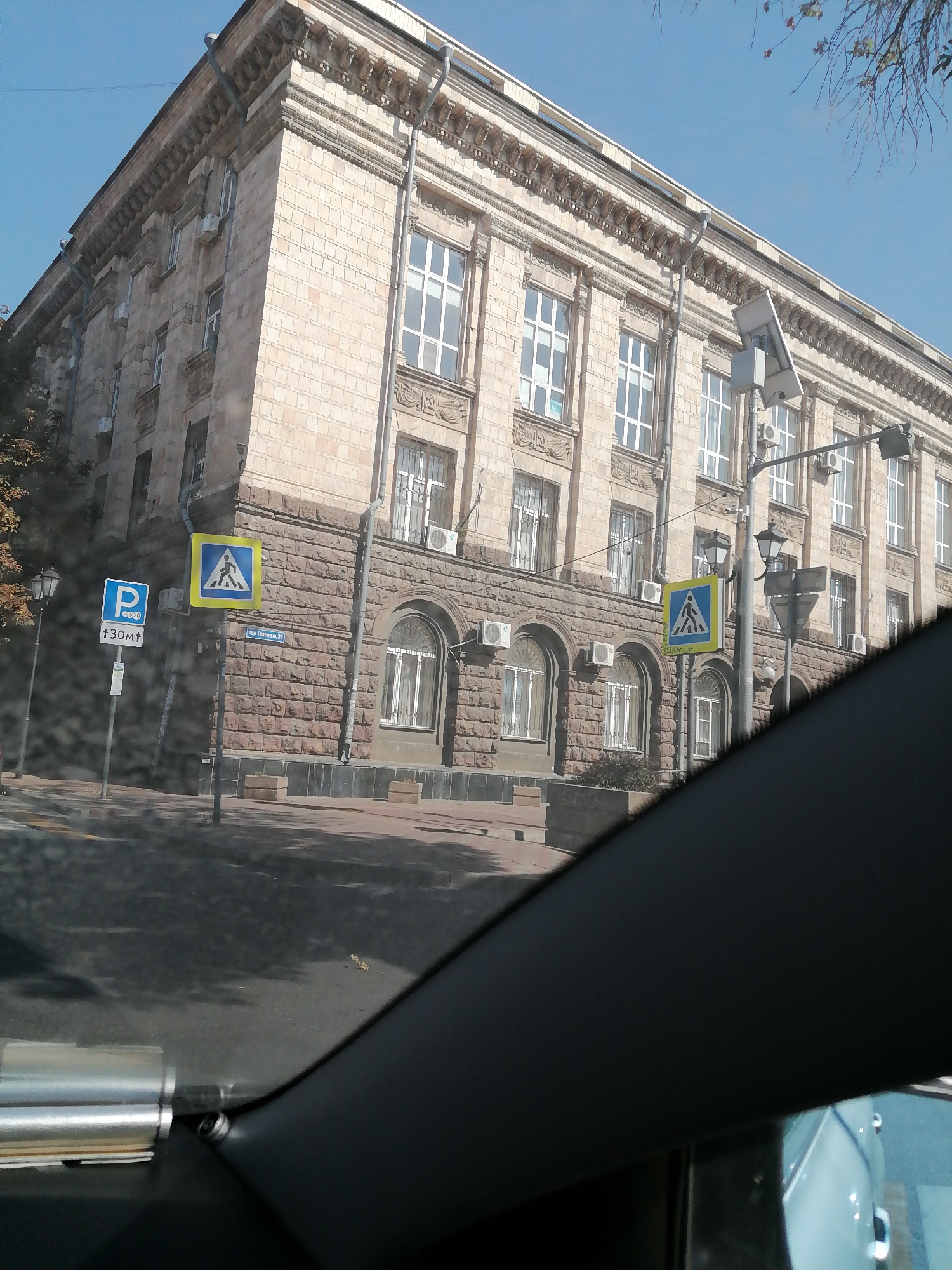 This is a daytime, slightly angled, exterior shot from inside a vehicle, looking out at a street scene in Rostov-on-Don, Russia. The primary subject is a large, stone-faced, multi-story building with numerous windows. The building's architecture features decorative cornices and arched ground-floor windows. Adjacent to the building, on the sidewalk, are two traffic signs: one is a blue square with a white "P" indicating parking and an upward arrow with "30m", and the other is a yellow square with a blue pedestrian crossing symbol. A street lamp is visible near the parking sign. The street in front of the building is paved, with shadows cast by trees on the road. A pedestrian crossing is marked on the road. A portion of the vehicle's interior, including a dark-colored dashboard or window frame and a metallic element, is visible in the foreground, obscuring part of the scene. The sky is clear and blue. The time of day appears to be mid-morning or afternoon, given the sunlight and shadows.