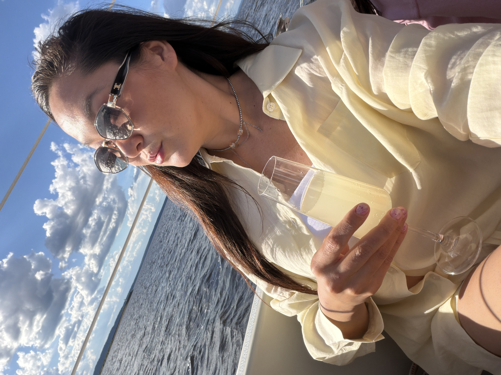A young woman is pictured in an outdoor setting, possibly on a boat or near a body of water under a partly cloudy sky. The location is Chaikovsky, Russia. She wears sunglasses and a light yellow blouse. In her right hand, she holds a glass of a light-colored beverage. She also has a delicate silver necklace with gems. The scene is brightly lit, suggesting it's daytime. The water surface is visible in the background, and the overall atmosphere appears relaxed and leisurely.