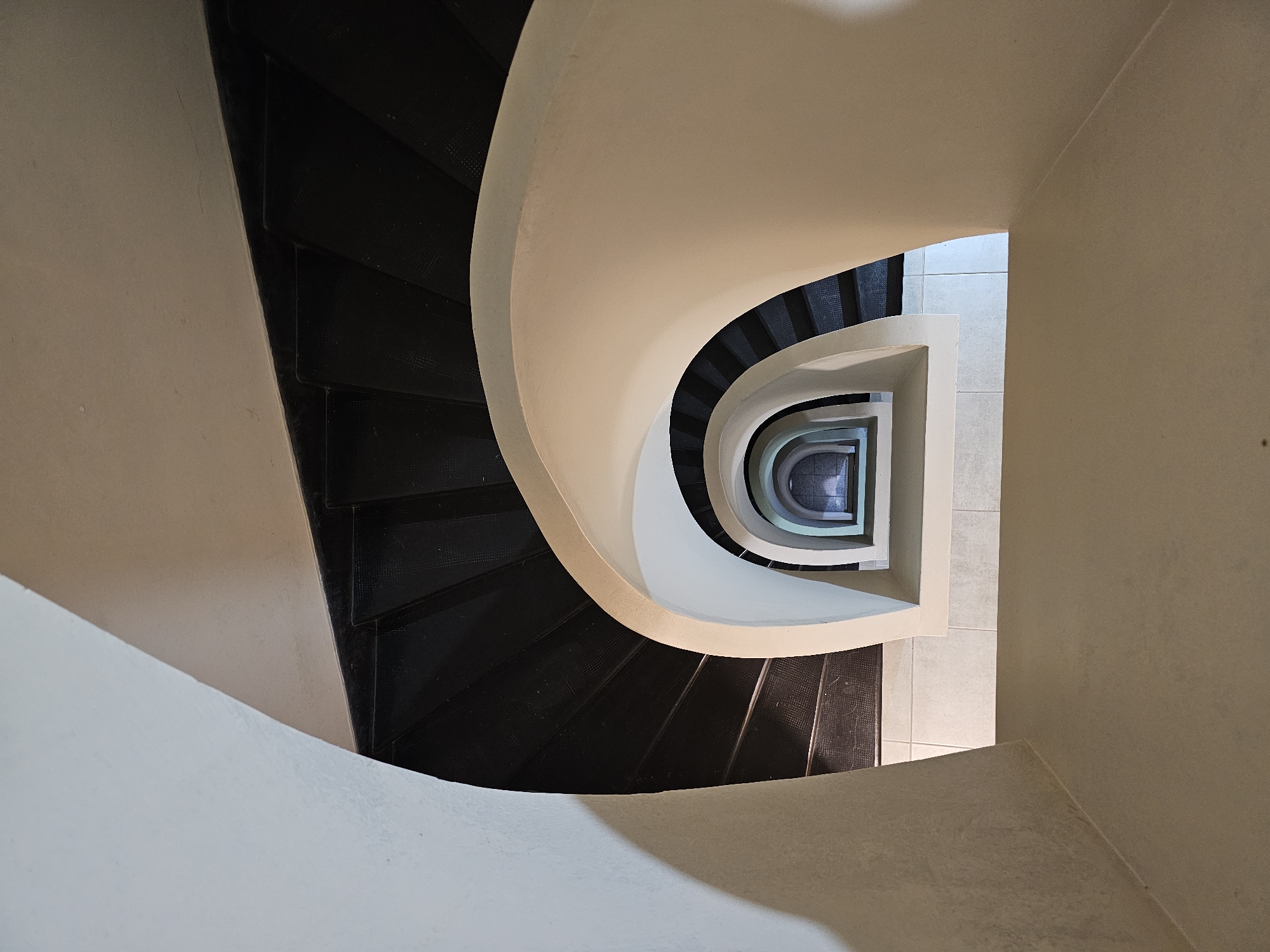 An overhead view looking down into a spiral staircase with dark treads and a light-colored wall. The staircase wraps around a central square space, with light-colored tile flooring visible at the bottom. There are no people in the frame. The location is not discernible based on the visual information provided.