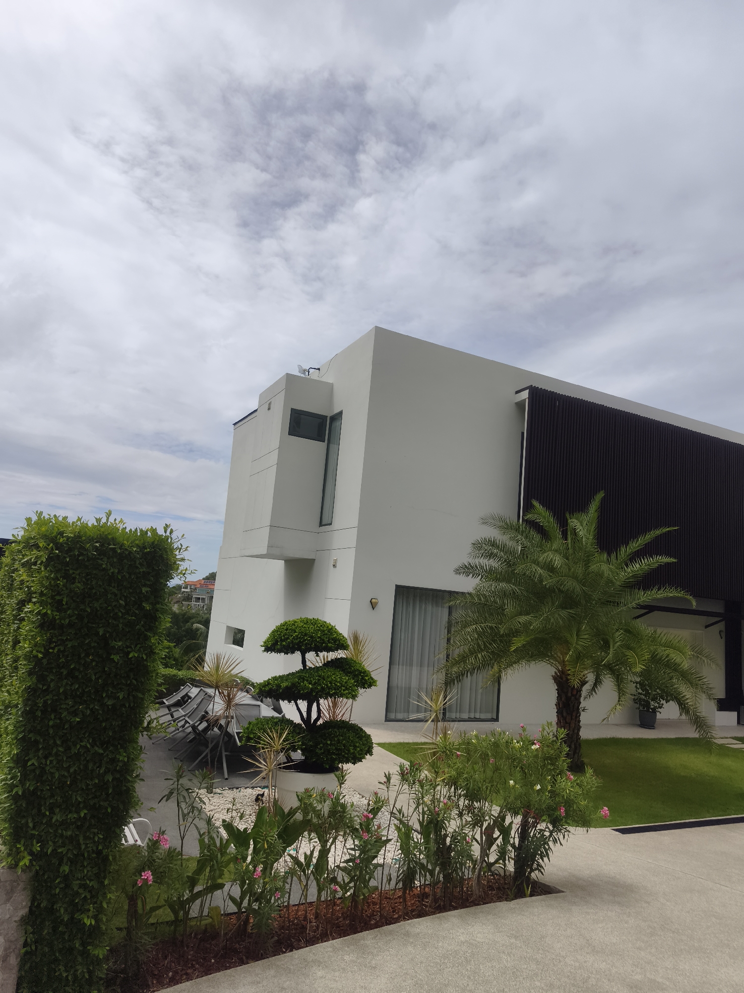 The image displays a modern architectural building nestled within a tropical landscape under an overcast sky. The main subject is a sleek, multi-story white building, likely a private residence or resort, characterized by clean lines and sharp angles. A distinct architectural element on the left side of the building protrudes outward, featuring a tall, narrow window. The upper right portion of the building boasts a dark, slatted facade that provides a strong contrast to the white walls. A large glass door or window is visible on the ground floor, partially obscured by plants.
The foreground and surrounding areas are richly landscaped. A tall, dense green hedge frames the left side of the scene. Near the building's base, a meticulously shaped, multi-tiered topiary tree stands out. A vibrant palm tree with full green fronds grows prominently in front of the building, and another smaller palm is visible further right. A flower bed in the lower foreground showcases pink and white blossoms amidst lush green foliage, mulched at its base. Patches of neatly manicured green grass are visible adjacent to the building and along the light-colored concrete pathways. Behind the hedge on the left, distant buildings and green hills can be seen.
The sky is heavily clouded and grey, indicating an overcast daytime. No specific activities or people are depicted, creating a peaceful and static scene. The architecture and tropical vegetation are consistent with the provided location context of Baan Tai, Thailand. There is no visible text in the image.