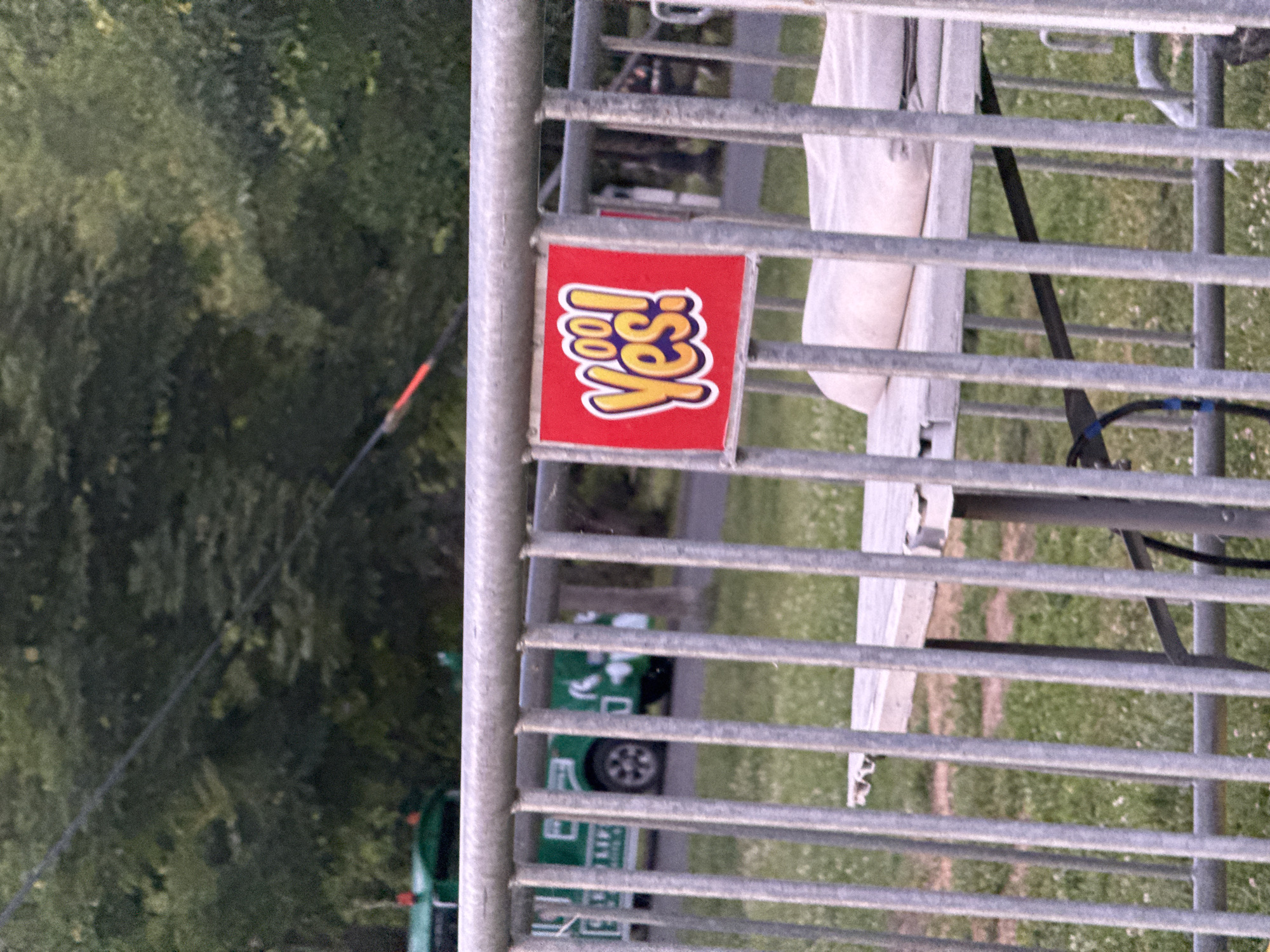The image captures a close-up of a metal barricade in Burgas, Bulgaria. Attached to the barricade is a bright red sign with a cartoonish yellow and white "Yes!" inscription. Behind the barricade, a green and white vehicle, possibly a small truck or utility vehicle, is partially visible on a paved surface. The background consists of lush green trees, suggesting an outdoor, possibly park-like setting. The lighting indicates it is daytime.