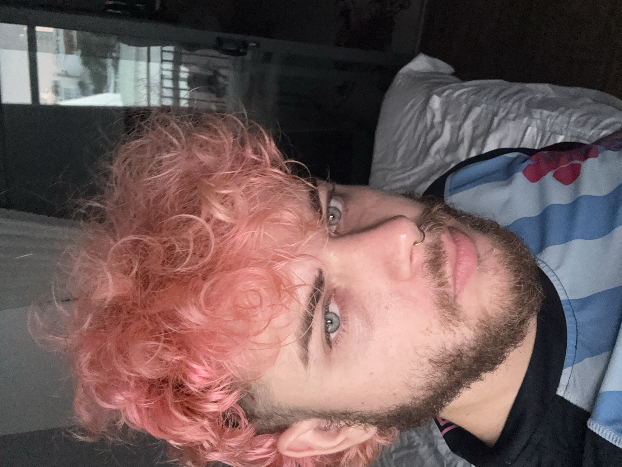A close-up, rotated selfie captures a person with distinctive features, likely taken indoors in Đà Nẵng, Vietnam. The subject, who appears to be male, has fair skin, light blue or green eyes, and thick, dark eyebrows. His hair is a vibrant, voluminous pink and styled in tight curls. He has a neatly trimmed beard and mustache, and a small golden-toned hoop nose ring adorns his right nostril.
The individual is positioned as if lying down, with his head tilted, looking directly at the camera. He is wearing a blue and white striped shirt with an additional pink/red abstract or floral pattern. A dark-colored collar or garment is visible underneath the striped shirt. Behind his head and upper body, there's a white, crumpled pillow or bedding, reinforcing the indoor, relaxed setting, possibly a bedroom. In the background, to the left, a window or reflective surface reveals glimpses of outdoor structures or buildings, suggesting an urban environment. The lighting is soft, creating a slightly subdued atmosphere. No discernible text is visible in the image.