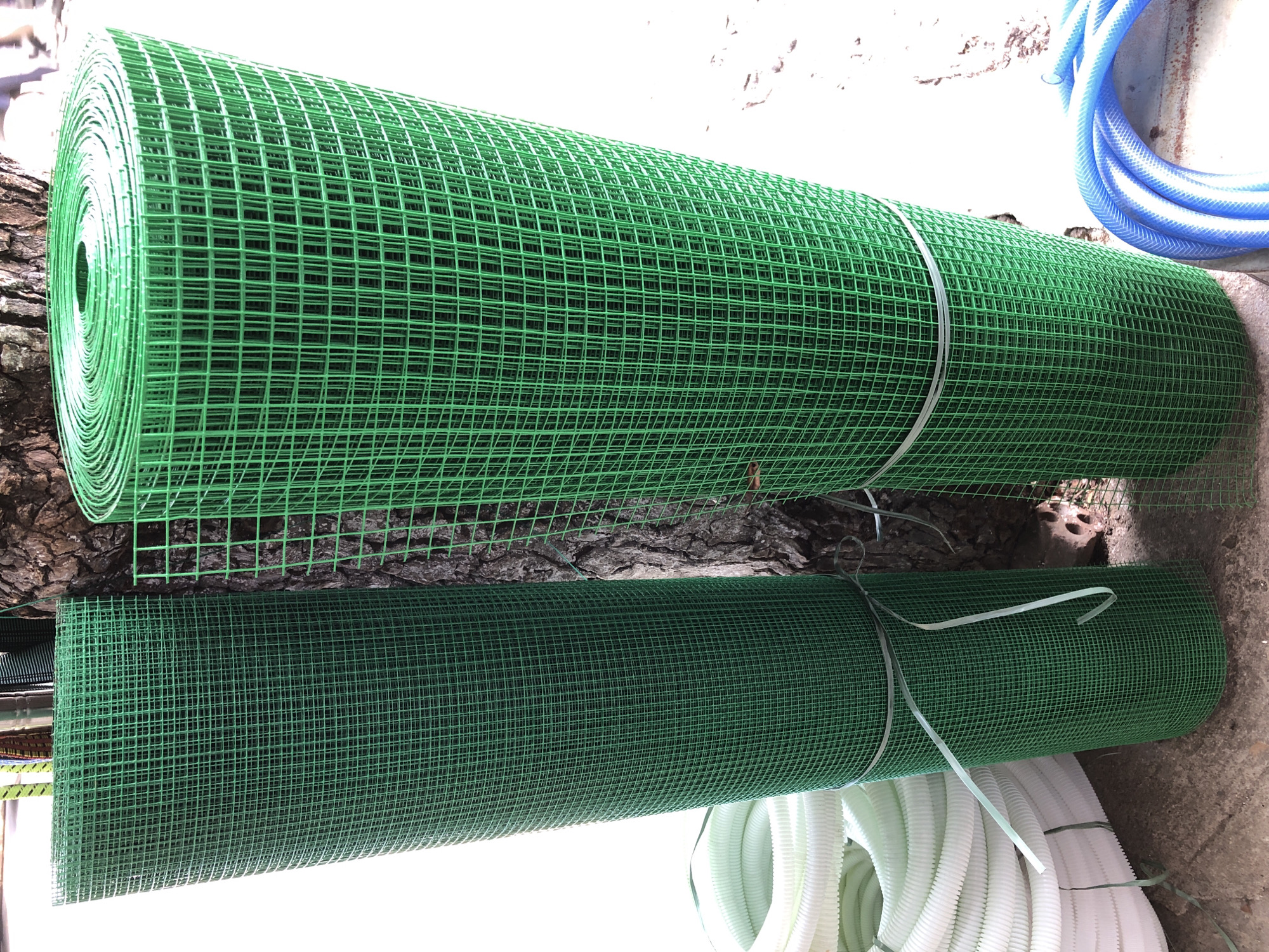 Two rolls of green plastic mesh fencing are leaning against a tree. One roll is on the left, and the other is on the right, with the tree trunk in between them. A stack of white corrugated tubing is visible to the left of the left roll. A blue hose is coiled on the ground to the right of the right roll. The ground appears to be concrete or dirt. The scene is outdoors, with a bright, white background that suggests a sunny day. The overall impression is of a commercial setting, likely a hardware store or garden supply business. The location is the city of Da Nang, Vietnam.