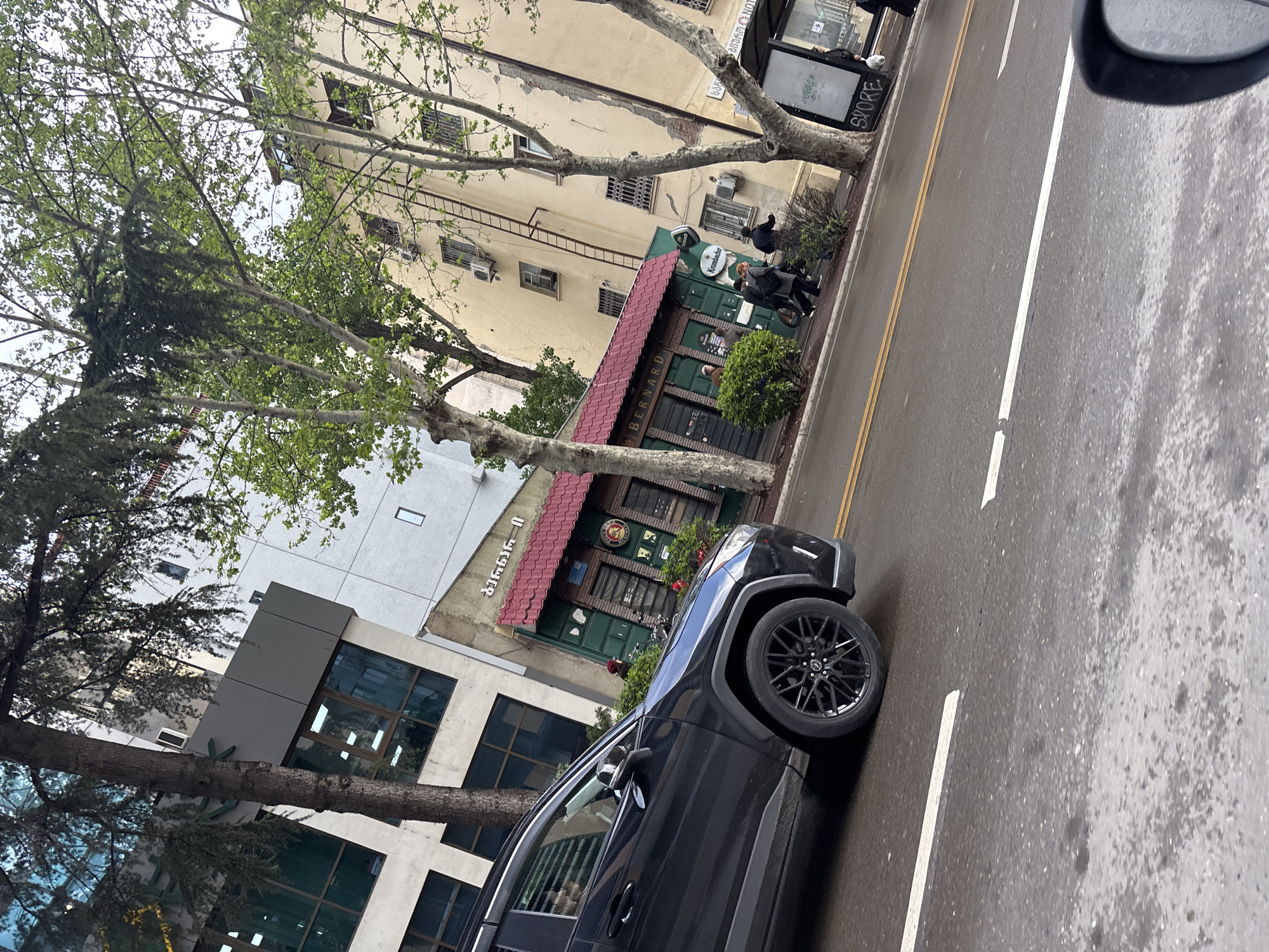 A street-level, medium-shot photograph taken from a vehicle, facing a street in Tbilisi, Georgia, shows buildings, trees, people, and a car. A black SUV is partially visible on the right side of the frame. The road surface appears wet. A beige building is present, with signs of weathering. A two-story building with a red awning and green facade is visible; the awning bears the name "Bernard." A sign in Georgian script is affixed to a white building above the bar. Two pedestrians are on the sidewalk, near a bus shelter. A tree is present.