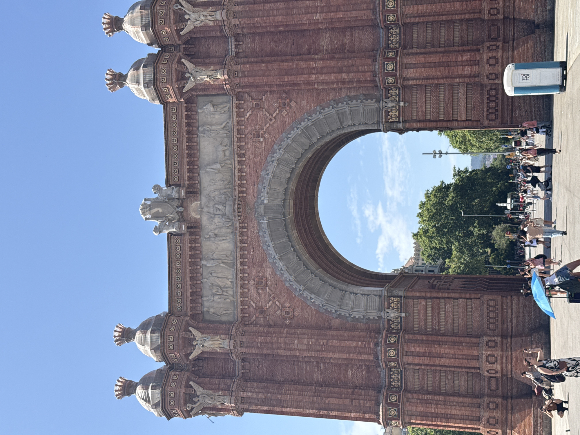The image shows the Arc de Triomf, a triumphal arch in Barcelona, Spain, on a bright, sunny day with a clear blue sky and scattered clouds. The arch is made of red brick and features intricate carvings and sculptures, including winged figures and reliefs depicting historical scenes. The archway itself is large and semicircular.
In front of the arch, there is a paved plaza with a considerable number of people walking, standing, and taking photographs. Among the crowd, some individuals are holding up their phones to capture images. A bright blue umbrella is visible near the center of the frame, offering shade to someone.
To the right of the arch, a portable toilet is situated on the pavement. The background reveals lush green trees and some buildings, suggesting an urban setting within the city. The overall atmosphere is that of a popular tourist attraction on a busy day.