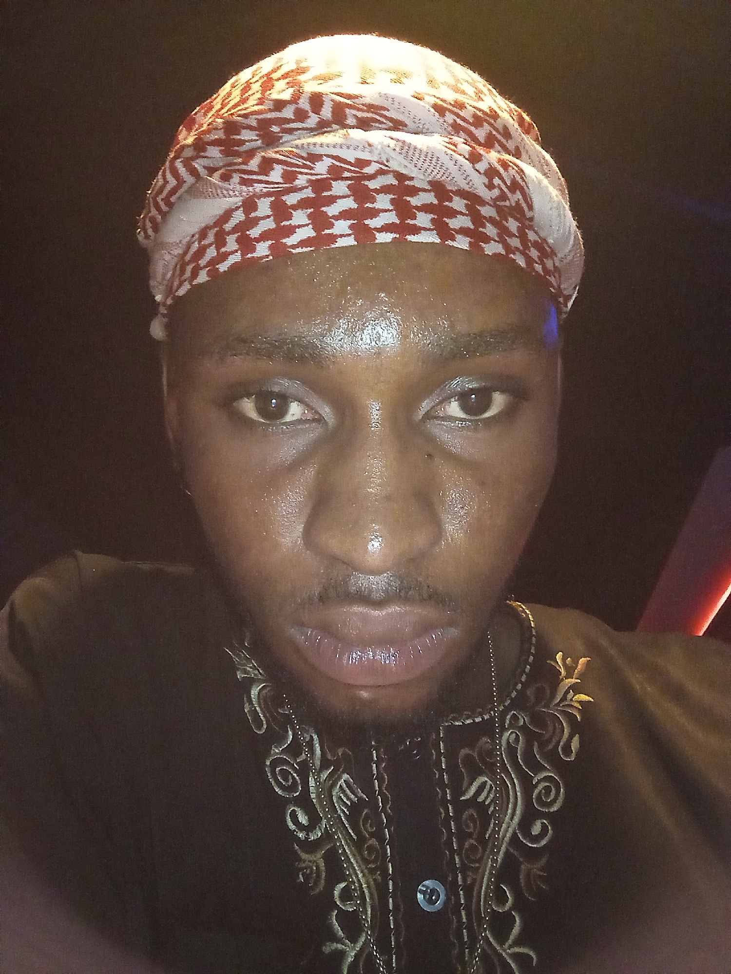 A close-up, dimly lit photograph shows a dark-skinned man wearing a red and white patterned headdress. He is also wearing a dark garment with ornate gold-colored embroidery around the collar. His face appears to be glistening with sweat or moisture. A thin chain necklace is visible around his neck. The background is dark. The city and location are unknown.