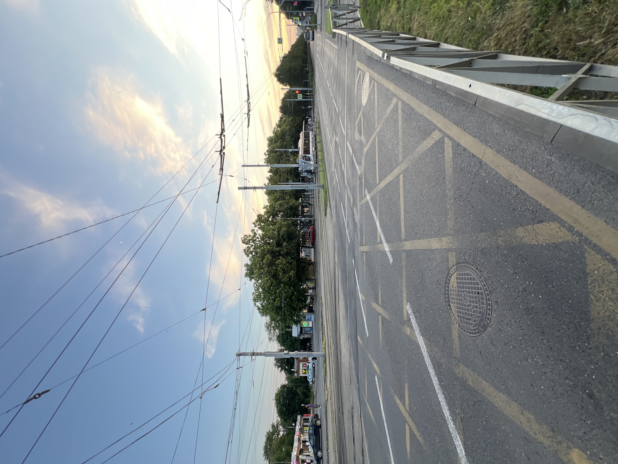 The image shows a street intersection in Krasnodar, Russia, at dusk. The sky is a mix of blue and pink, with scattered clouds. Overhead, a complex network of trolleybus wires crisscrosses the frame. The road is asphalt with yellow and white road markings, including what appear to be waiting zones or bus stops. A manhole cover is visible on the road.
On the left side of the road, there are several buildings that appear to be shops or kiosks, with people walking in front of them. A black car is parked on the side of the road. Further down the street, a light blue car is visible. Trees line the sidewalks and median.
In the middle of the intersection, a trolleybus is stopped. Across the intersection, more trees and buildings are visible. Traffic lights are present, one of which is red. A white bus is traveling away from the camera on the right side of the road. A metal fence runs along the right side of the foreground. The overall scene suggests a typical urban environment during the evening hours.