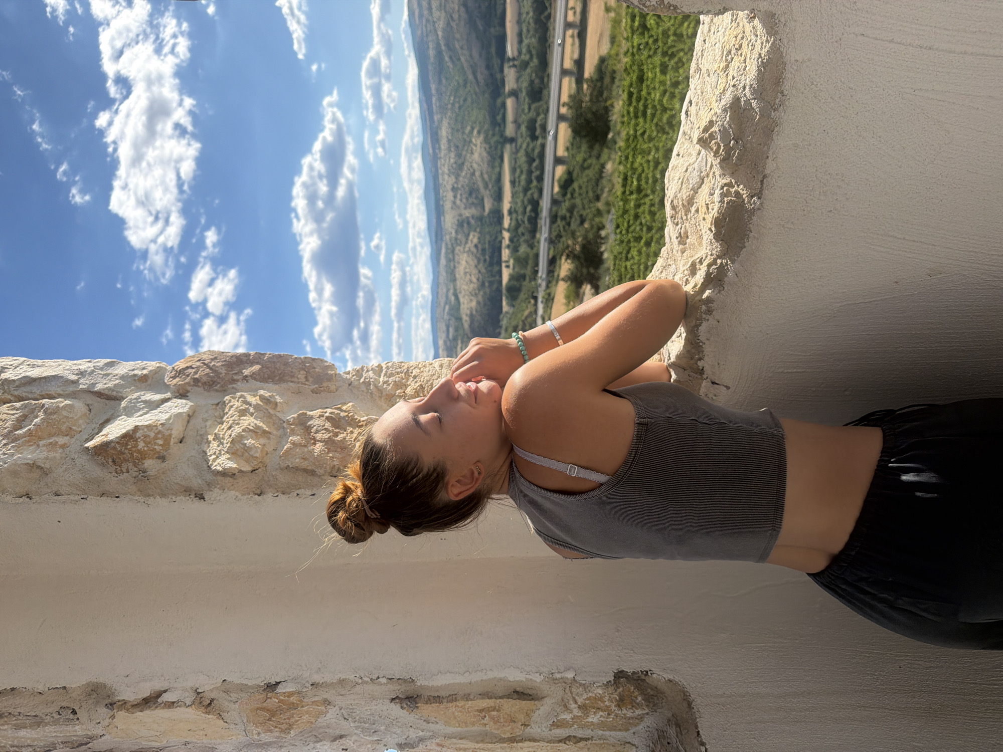 A young woman with her hair in a bun leans on a stone windowsill, looking out at a scenic view. She wears a dark grey tank top and dark pants. Her arms are crossed, and her hands are touching her face. Her eyes are closed, and her face is tilted upwards, suggesting she is enjoying the sunlight. The background shows a valley with green vineyards and a highway with cars passing by. The sky is blue with scattered white clouds. The stonework of the windowsill and the surrounding wall is rough and textured. The lighting suggests it is daytime, with the sun casting shadows. The location is the town of Kocherinovo, Bulgaria, specifically a viewpoint within the town.