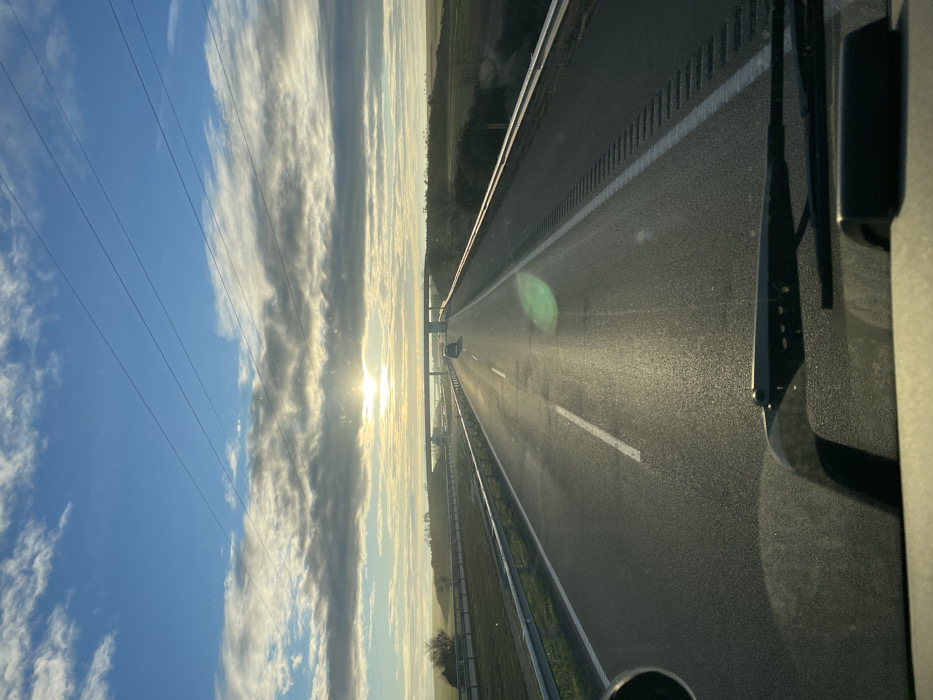 A sun-drenched highway stretches into the distance, its asphalt glistening. Overhead power lines run parallel with a dramatic sky featuring blue patches and long, striated clouds. A vehicle's windshield wiper is visible in the corner. Guardrails flank the road, and grassy strips extend beyond. Another car appears far ahead on the highway. The scene is taken from inside a vehicle.