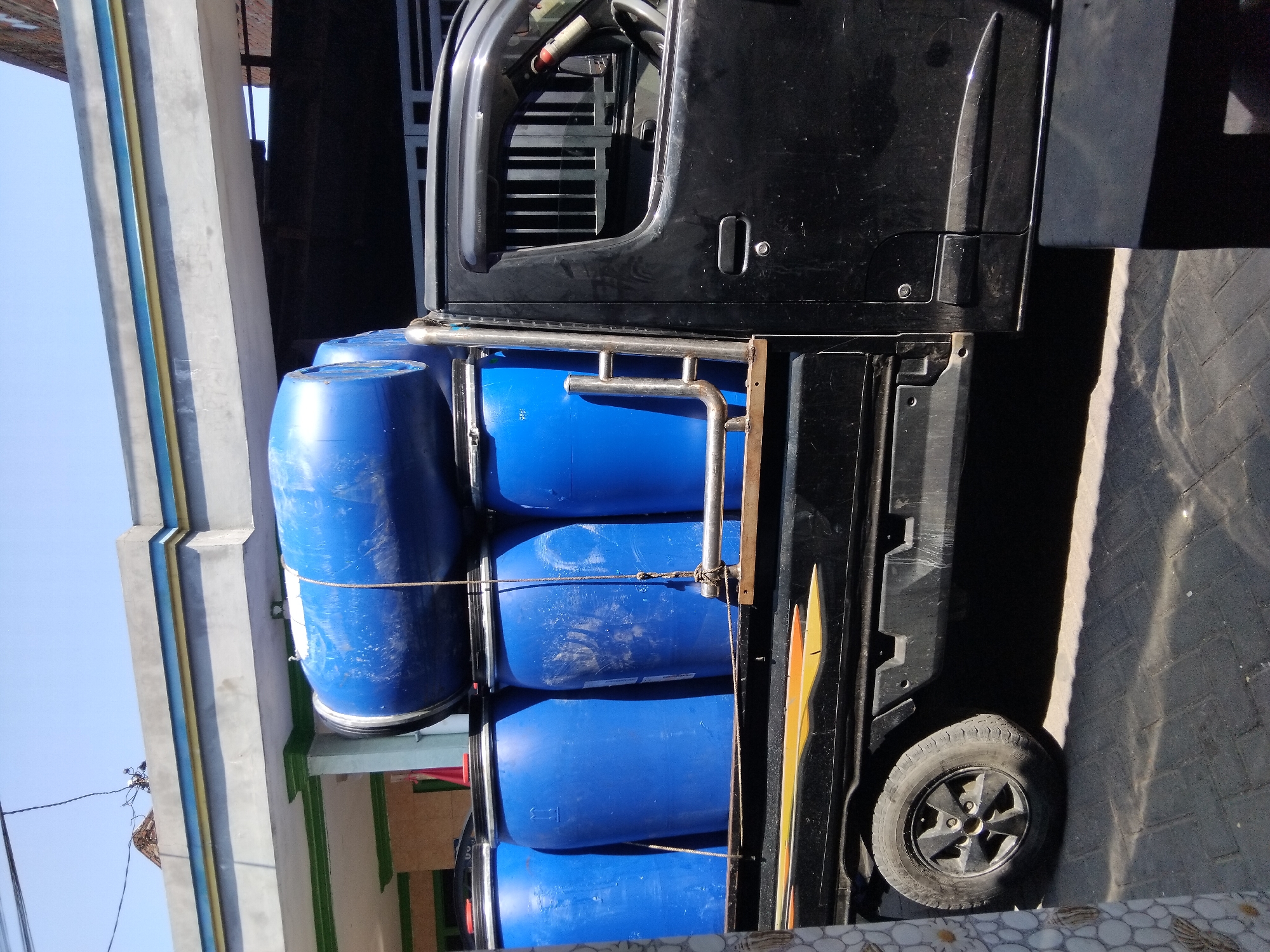 A black pick-up truck is parked on a paved surface under a clear blue sky. The truck bed is filled with multiple large, blue plastic barrels, some of which are stacked. One barrel is positioned horizontally on top of others, secured by ropes. The truck has a silver railing along the side of the bed. A residential building with a grey, blue, and yellow striped awning is visible in the background, along with a section of a dark-colored fence. The image appears to be taken in the city of Kepuh Kiriman, Indonesia.