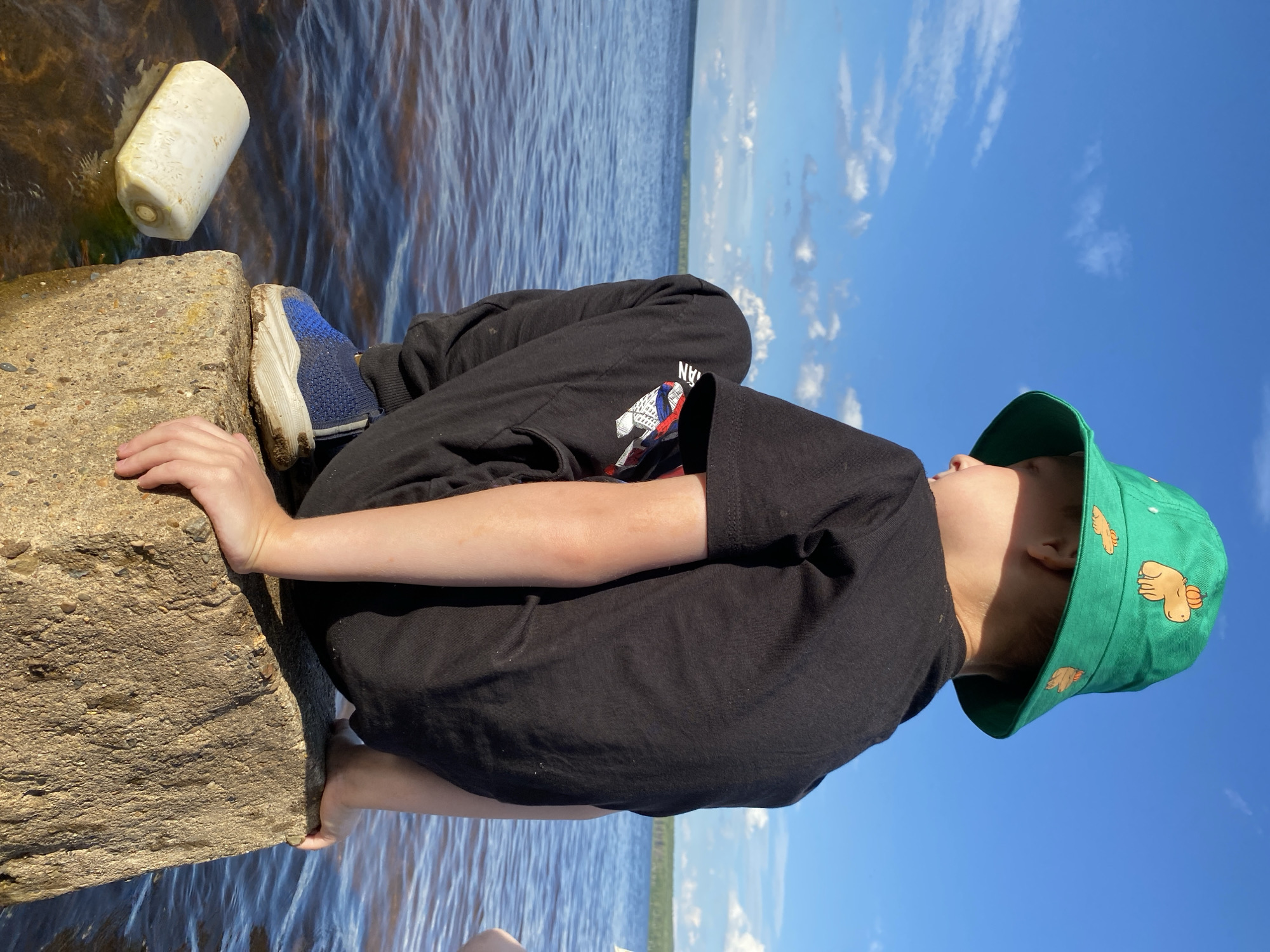 In Chaikovsky, Russia, a young person wearing a black t-shirt and black pants, along with a green bucket hat adorned with cartoon capybaras, sits on a concrete block at the edge of a body of water. The person is facing away from the camera, looking out over the water. A white plastic float is partially submerged in the water to the left of the concrete block. The sky is blue with scattered white clouds, and a distant treeline is visible across the water. The scene appears to be an outdoor, natural setting.