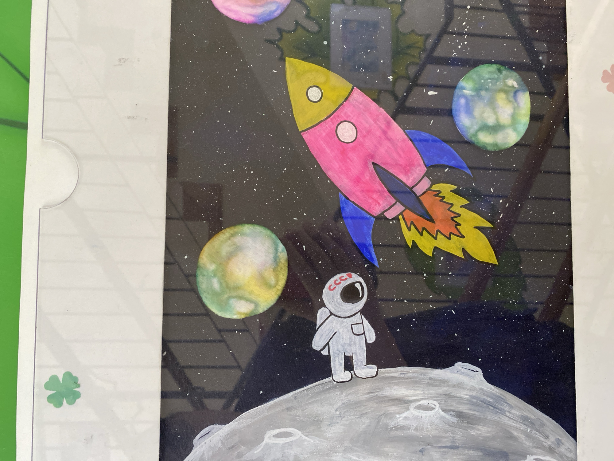 The image depicts a child's drawing of a space scene. A large gray moon-like object is on the left side of the frame. An astronaut figure, drawn in white with a red "CCCP" insignia on the helmet, is positioned near the moon. A colorful rocket, primarily pink with a yellow nose cone and blue fins, is shown in flight, with flames depicted in orange and yellow. Several planets, rendered in various colors and textures, are scattered throughout the dark, star-speckled background. A green four-leaf clover is visible in the upper left corner of the white border surrounding the drawing. The scene is framed within a white border. There are reflections on the surface of the image, obscuring some details. This artwork is located in the city of Kaliningrad, Russia.