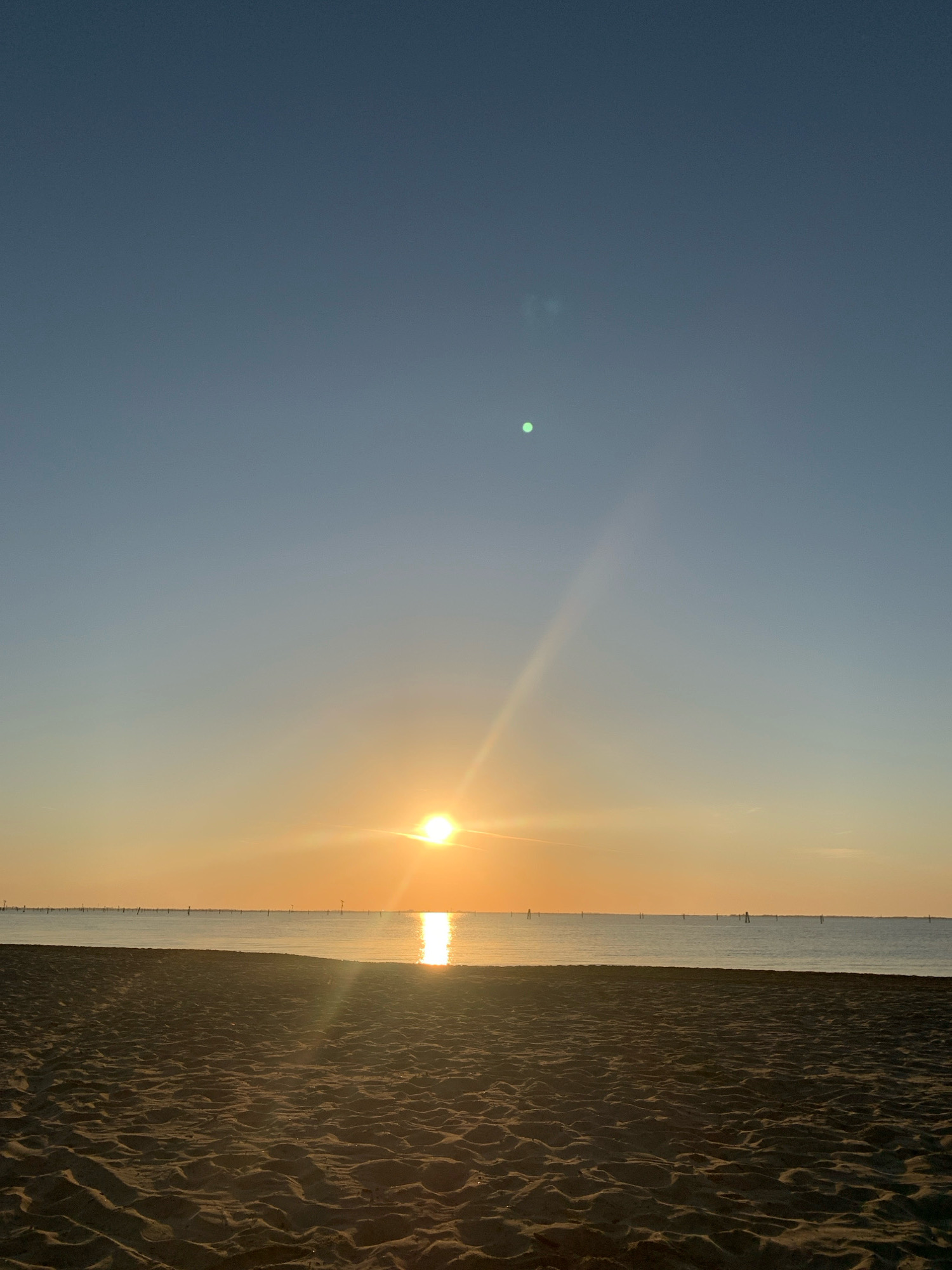 This image depicts a serene beach scene at sunrise, with the sun casting a warm glow over the tranquil sea. The foreground is dominated by the sandy texture of the beach, with subtle ripples and shadows indicating the play of light. In the distance, a thin line of buoys or markers stretches across the horizon of the calm water, suggesting a boundary or navigation aid. The sky transitions from a pale blue at the top to a soft orange hue near the horizon, where the sun is just beginning to rise, its rays reflecting brightly on the water's surface. The overall atmosphere is peaceful and tranquil, characteristic of a quiet dawn by the sea. This scene is likely located on the coast of Comacchio, Italy, a region known for its extensive lagoons and sandy shores, offering such picturesque sunrise views over the Adriatic Sea.