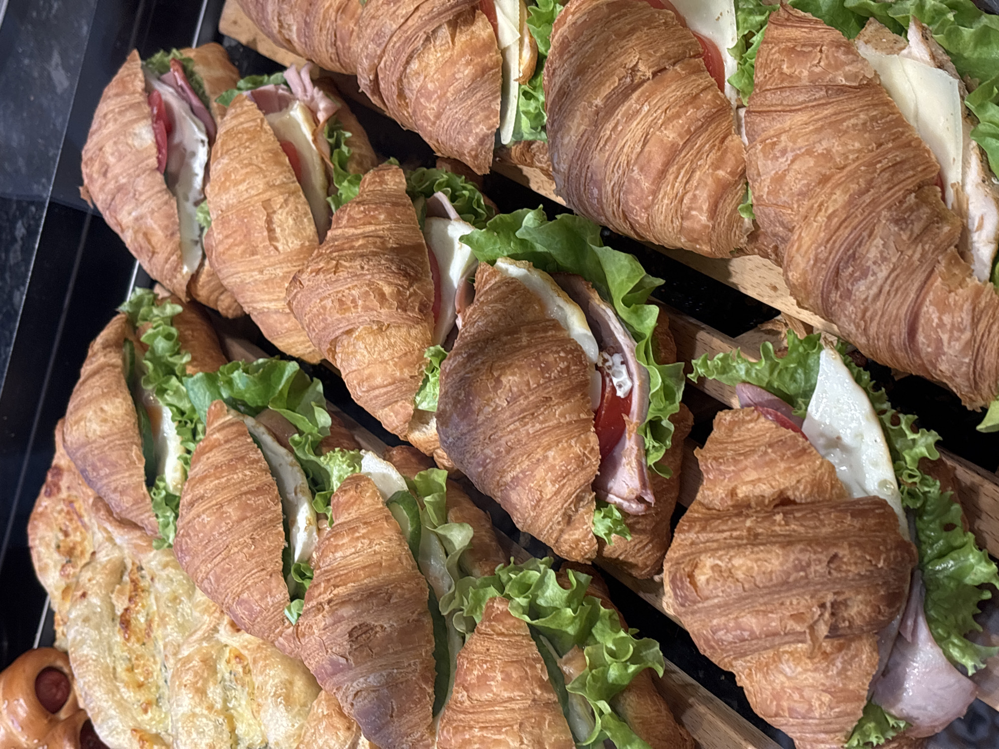 A close-up, overhead shot showcases a variety of croissant sandwiches and pastries arranged on wooden boards. The croissants are golden brown and flaky, with fillings visible. Some croissants contain lettuce, tomato, cheese, and what appears to be ham or turkey. Other pastries, including what seems to be a sausage roll and a large, layered pastry with cheese, are also present. The background is dark, suggesting an indoor setting, possibly a bakery or cafe. The image does not provide enough information to determine a specific location in Nizhny Novgorod, Russia.