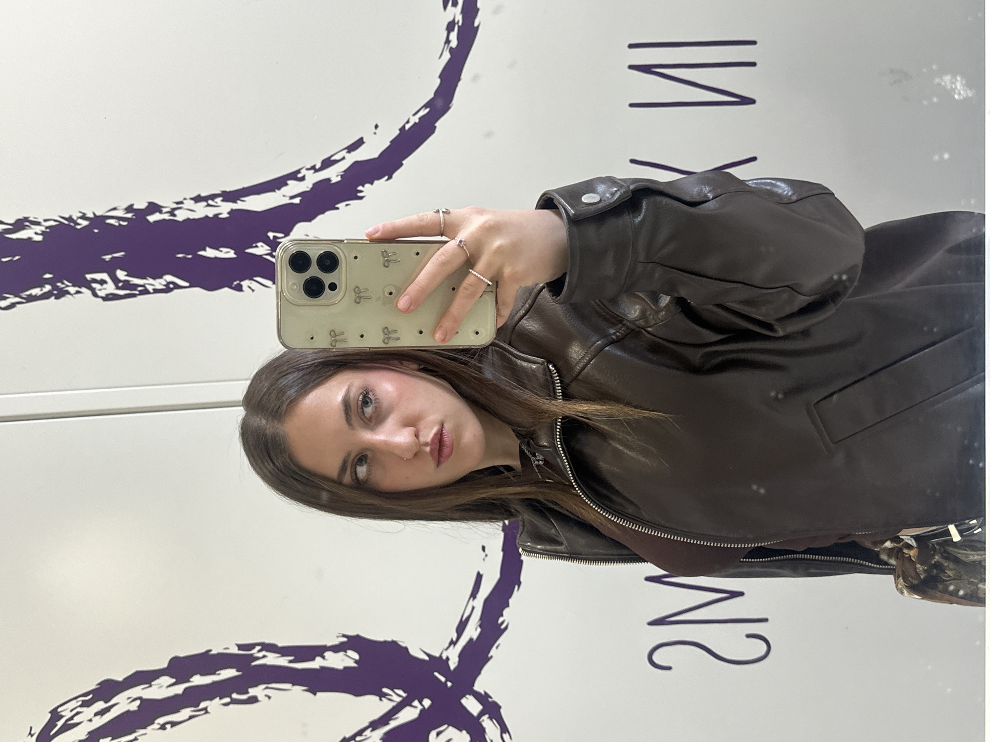 A young woman in a brown leather jacket is taking a selfie in front of a mirror with purple markings on it. She is holding up a phone with a light-colored case adorned with bows. She is wearing rings on her fingers. Based on the mirrored inscription “MX IN”, it appears she is in Mexico City, Mexico.