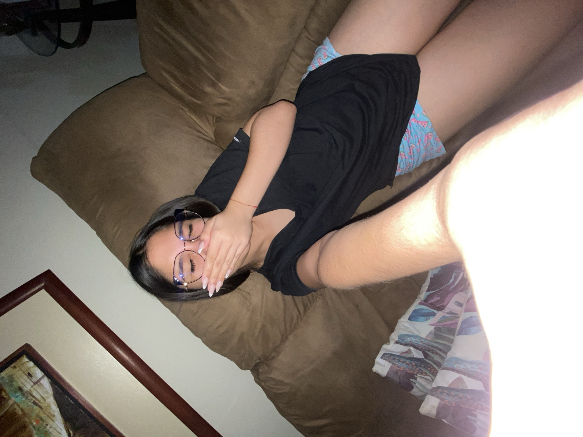 A young woman lies on a brown sofa, wearing a black shirt and patterned shorts. She is covering her mouth with her hand and wearing glasses. A framed painting is visible on the left. A brightly lit object or area is in the lower right. There is no contextual information or element in the image that allows inferring the location of the scene to be in the city of Cotoperíz, Venezuela.