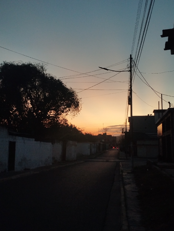 This image depicts an urban street scene in Guacara, Venezuela, during twilight, with the sky transitioning from light blue overhead to an orange-yellow hue near the horizon. The foreground features a dark asphalt road with a curb and sidewalk on the right. Along the left side of the street, a sequence of single-story structures with light-colored, possibly weathered walls and dark, rectangular door openings are visible, partially obscured by a large, dark-silhouetted tree. On the right, a prominent utility pole supports multiple overhead wires and a streetlamp fixture, angled to illuminate the road. The street extends into the distance, flanked by further buildings of varying heights, some appearing under construction or in disrepair. Faint red lights, consistent with vehicle taillights, are discernible in the far background. No individuals or specific actions are visible within the frame.