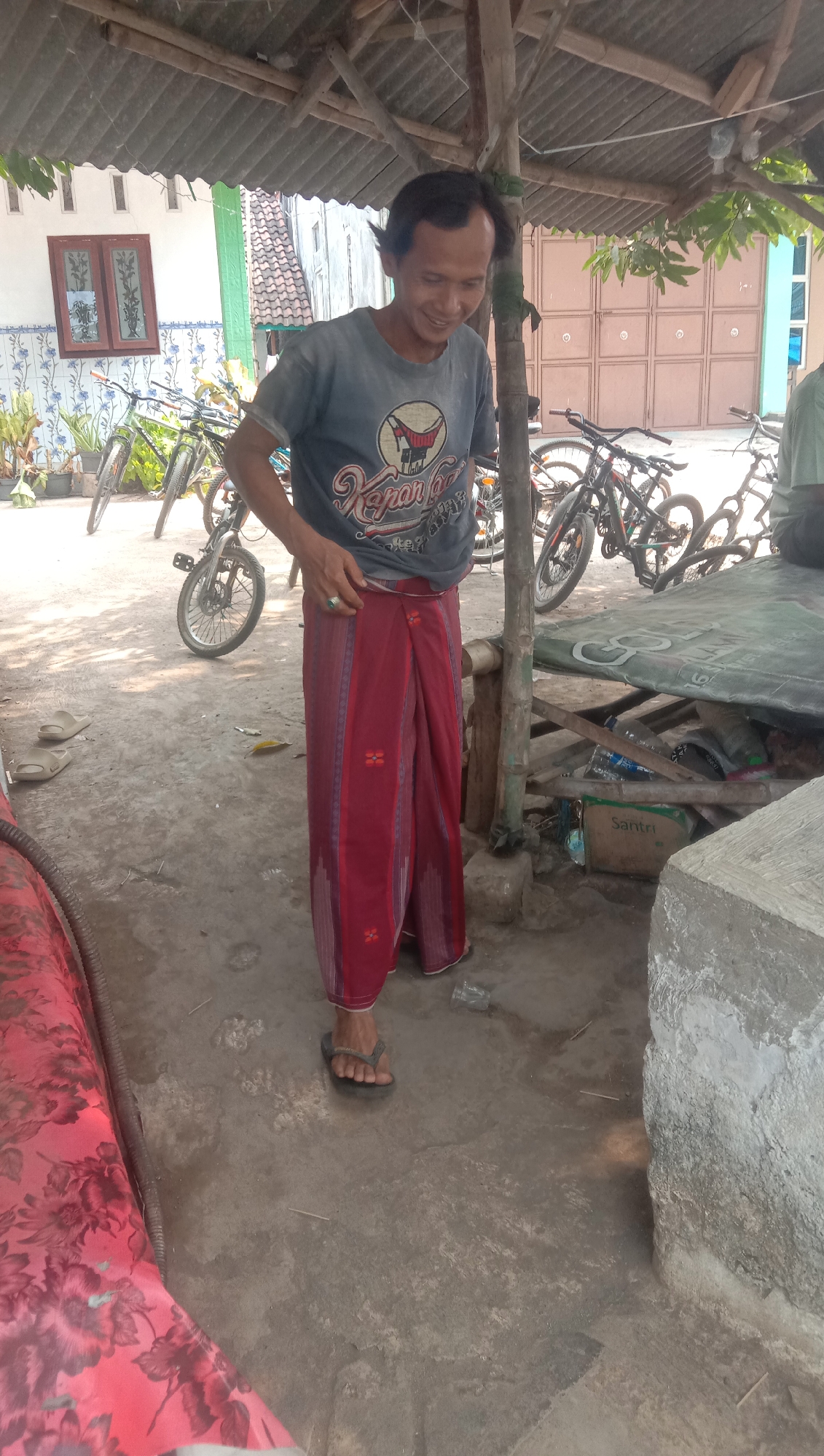In Ngempit, Indonesia, a man stands under a rustic, open-air shelter with a corrugated metal roof supported by wooden beams. He wears a dark grey t-shirt with a graphic design and a red sarong with a repeating pattern of red floral motifs. His right hand is raised, adjusting the sarong. He wears dark flip-flops. In the background, several bicycles are parked. To the left, a white building with a dark-framed window and decorative blue and white tile accents is visible. The ground is a dirt or concrete surface. A table covered with a green tarp, containing bottles and boxes, is to the right of the man. A large concrete block is also present in the foreground.
