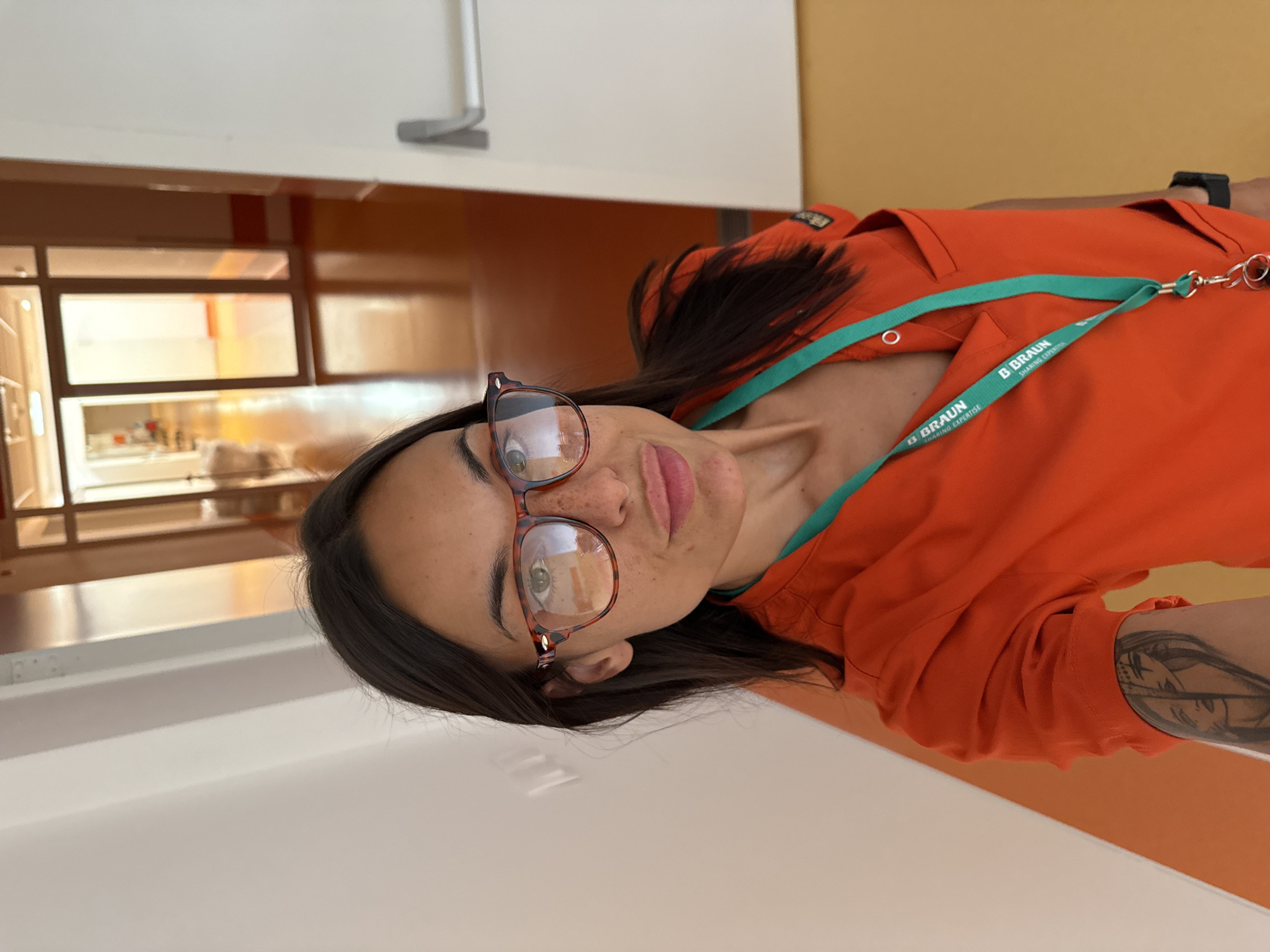 A medium shot photo shows a young woman wearing an orange scrub top and glasses. She is standing in a hallway that appears to be in a medical facility. The hallway has white walls, orange floors, and glass doors in the background. The woman is looking at the camera and smiling. There is a tattoo on her left arm. A lanyard with a logo that says "B. Braun" is around her neck. The woman is positioned in the foreground, and the hallway recedes into the background.