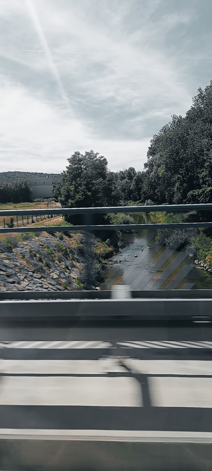 The image captures a scene from a bridge overlooking a river and surrounding landscape, likely taken from a moving vehicle due to motion blur in the foreground. In the foreground, two parallel grey railings run horizontally across the frame, with the lower railing exhibiting motion blur. Below the railings, the blurred surface of a road or pathway is visible, marked with dark and light parallel lines, suggesting speed.
The middle ground features a shallow, meandering river with rocky banks on the left. The river's surface reflects the sky and surrounding trees. On the left bank, a gently sloping area is covered with stones and dry vegetation. Behind the riverbank, a row of metal bollards with a fence separates the area from a field with sparse vegetation and a large, rectangular building with a flat roof, possibly an industrial or commercial structure, situated in the distance. A dense line of trees forms the backdrop, with taller trees to the right and a hill covered in vegetation further in the distance on the left. The sky is overcast with a bright ray of sunlight piercing through from the upper left, creating a slightly ethereal glow. A faint contrail from an aircraft is visible in the sky.
The location identified is Winterbach, Germany. The scene depicts a natural landscape integrated with infrastructure, common in suburban or semi-rural areas of Germany. The time of day appears to be daytime, possibly late morning or early afternoon, given the brightness of the sun. No people are visible in the image, and the focus is on the environment and the man-made structures within it.
