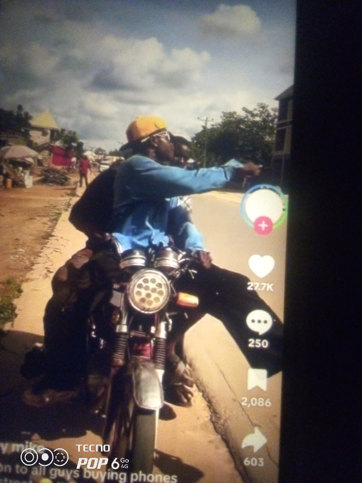 The image depicts a scene with two individuals on a motorcycle. The person in the front, wearing a yellow cap and sunglasses, is pointing to the right. They are dressed in a blue long-sleeved shirt. The second individual, seated behind them, is mostly obscured by the motorcycle. The motorcycle is a dark color with a prominent round headlight.
The environment appears to be an outdoor, possibly semi-urban or rural setting, with dirt roads and some buildings in the background. There are also some market stalls or makeshift structures visible. The lighting suggests it is daytime, with a cloudy sky. The image also contains on-screen interface elements, including icons for likes, comments, shares, and associated numbers (27.7K, 250, 2,086, 603), as well as text overlaying the bottom of the image, which includes "TECNOP POP 6 4G". The description of "Howayi, Nigeria" in the prompt seems to be an assumed location, as there are no explicit visual cues within the image to confirm this.