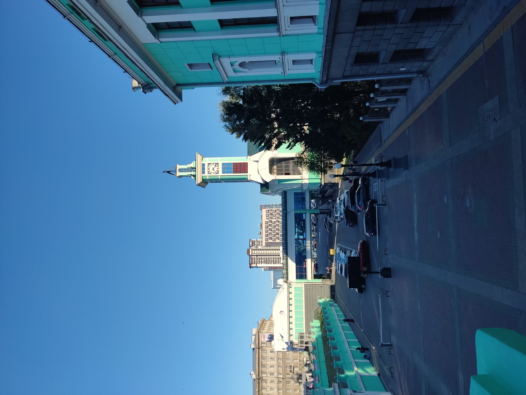 The image shows a view of the Novosibirsk-Glavny railway station in Novosibirsk, Russia, on a clear day. The foreground features a paved area with parked cars and a sidewalk. A bright teal colored building with a white trim and arched windows dominates the right side of the frame. In the mid-ground, a clock tower with a green roof and a white facade stands prominently, displaying a clock face and digital readouts for temperature. Beyond the clock tower, several other buildings of varying heights and architectural styles can be seen, including a modern high-rise building. The sky is a clear, bright blue with a few small dark specks that could be birds. Several cars are parked in designated areas, and a few pedestrians are visible. The overall scene depicts an urban environment with a mix of historical and modern architecture.