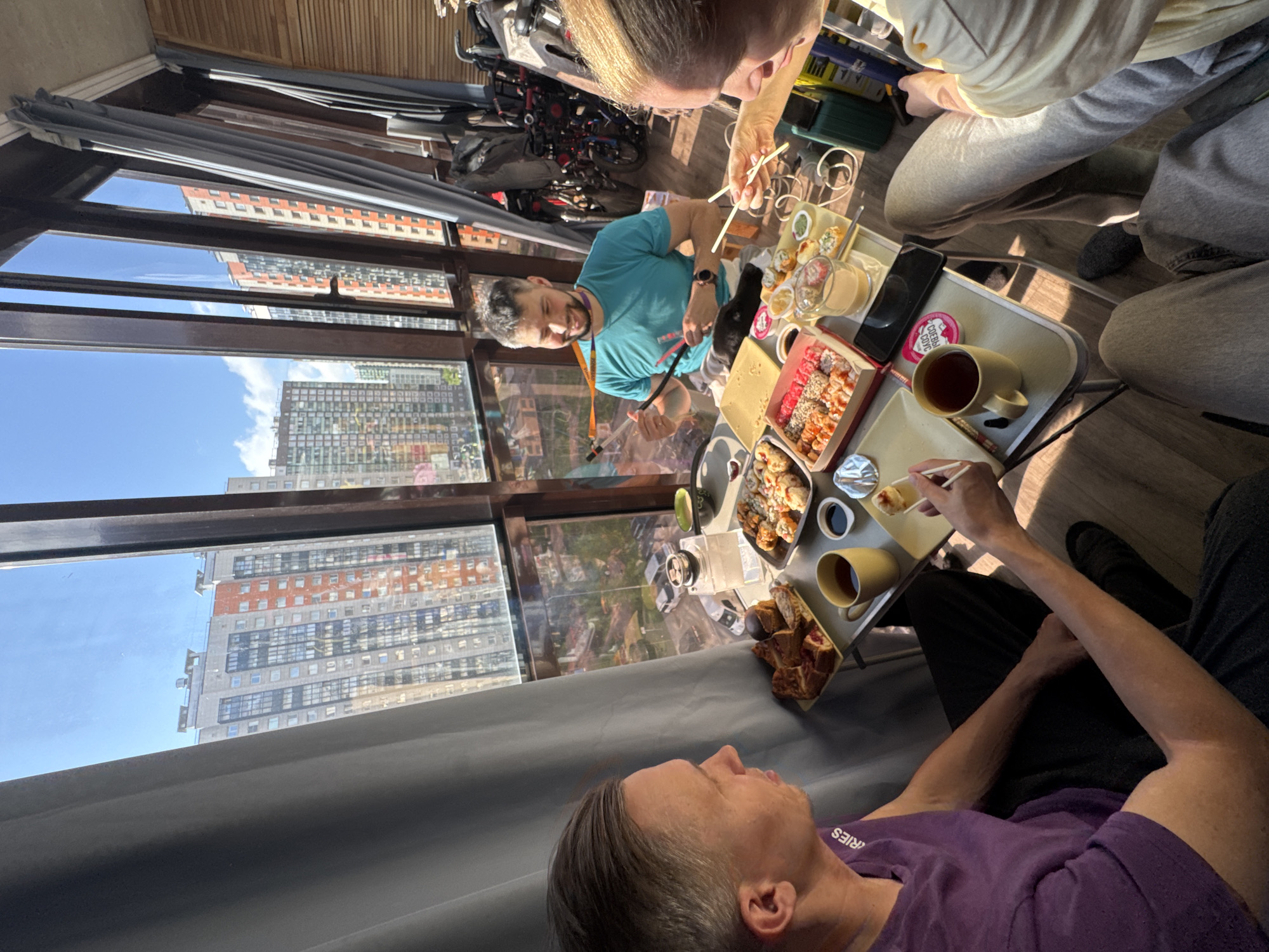 Here is a description of the image:
The image shows an elevated indoor scene, likely an apartment in Murino, Russia, as suggested by the cityscape visible through the large windows. Several individuals are positioned around a low portable table filled with sushi, small bowls, and drinks.
Three people are clearly visible and engaged in eating. A man wearing a purple shirt is lying down near the front of the frame. Another man in a light-colored shirt sits adjacent to the table. A third man wearing a turquoise shirt is picking up sushi with chopsticks.
The table holds the meal of sushi, containers, and cups. A cell phone is also on the table. Sunlight streams through the windows, illuminating the buildings outside and interior space. A wheelchair and assorted luggage are visible near the windows. The overall setting indicates a relaxed and informal meal being shared in an apartment overlooking the city.