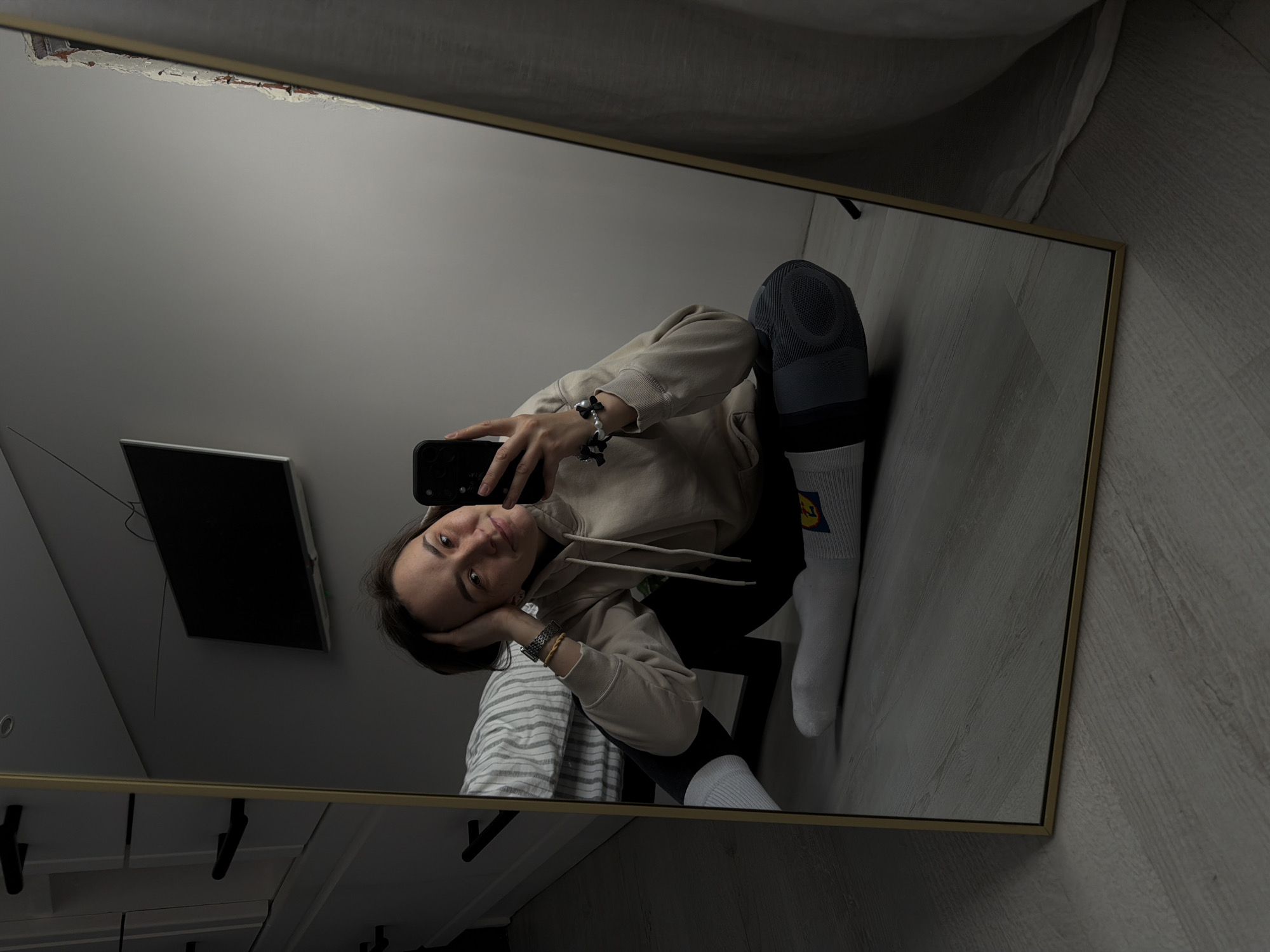 A young woman is taking a mirror selfie in an indoor setting. She is crouching and looking into her smartphone, which she is holding with her right hand. She is wearing a beige hoodie, black leggings, and white socks with a blue and yellow logo, possibly from Lidl. Her left hand rests on her head. Reflected in the mirror are a gray wall, a flat-screen television, part of a white striped garment, a white ceiling, and a light-colored wooden floor. The mirror has a thin gold frame. There is no verifiable indication in the image that the location is in the city of Chaikovsky, Russia.