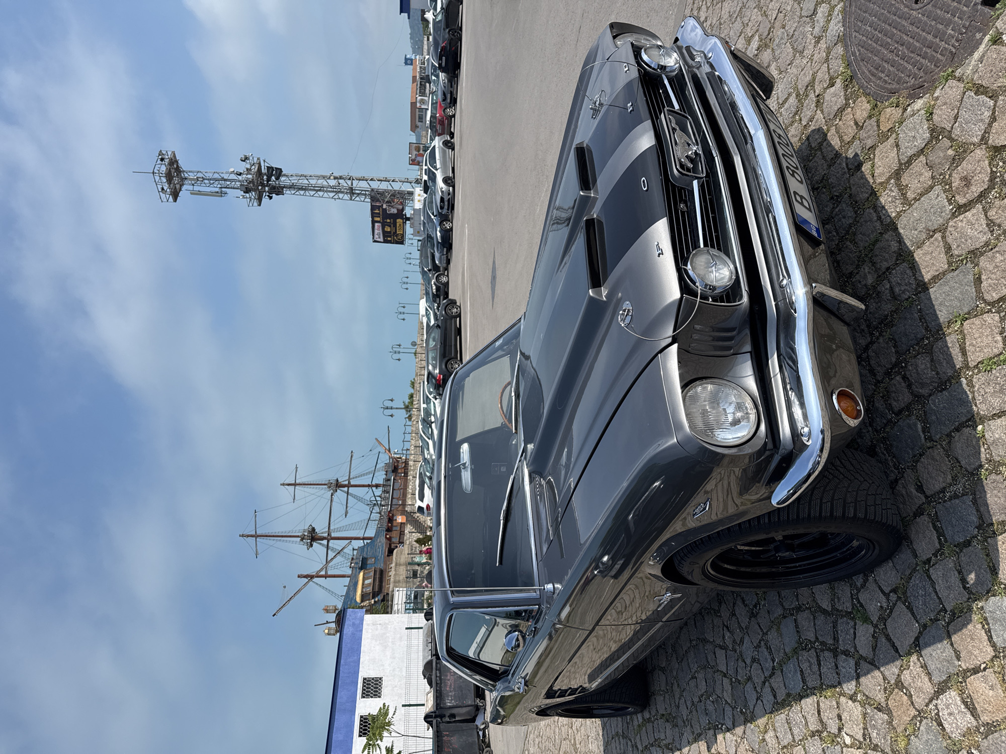 The image captures a classic grey Ford Mustang with dark grey racing stripes parked on a cobblestone surface, illuminated by bright daylight. The car is prominently featured in the foreground, angled towards the right, showcasing its chrome front bumper, grille with the Mustang horse emblem, circular headlights, and hood scoops. The Mustang has black multi-spoke wheels and a European-style license plate starting with "B 80000" on the front. Shadows from the car and other objects are visible on the cobblestones, indicating a sunny day.
In the mid-ground, behind the Mustang, a parking lot with several modern cars is visible. Further back and to the left, a large, intricate metal lattice structure, possibly a communication or observation tower, stands tall against the sky. A large black sign with white text, showing what appears to be "pizza" and a coffee cup icon, is attached to this structure.
To the right and further back, a tall-masted sailing ship, likely a historical replica or a moored attraction, is docked near some buildings. Its rigging and sails are visible against the blue sky with scattered white clouds. The overall setting appears to be a waterfront area or a large parking lot adjacent to a port or tourist destination. No people are visible in the scene. The weather is clear and bright.