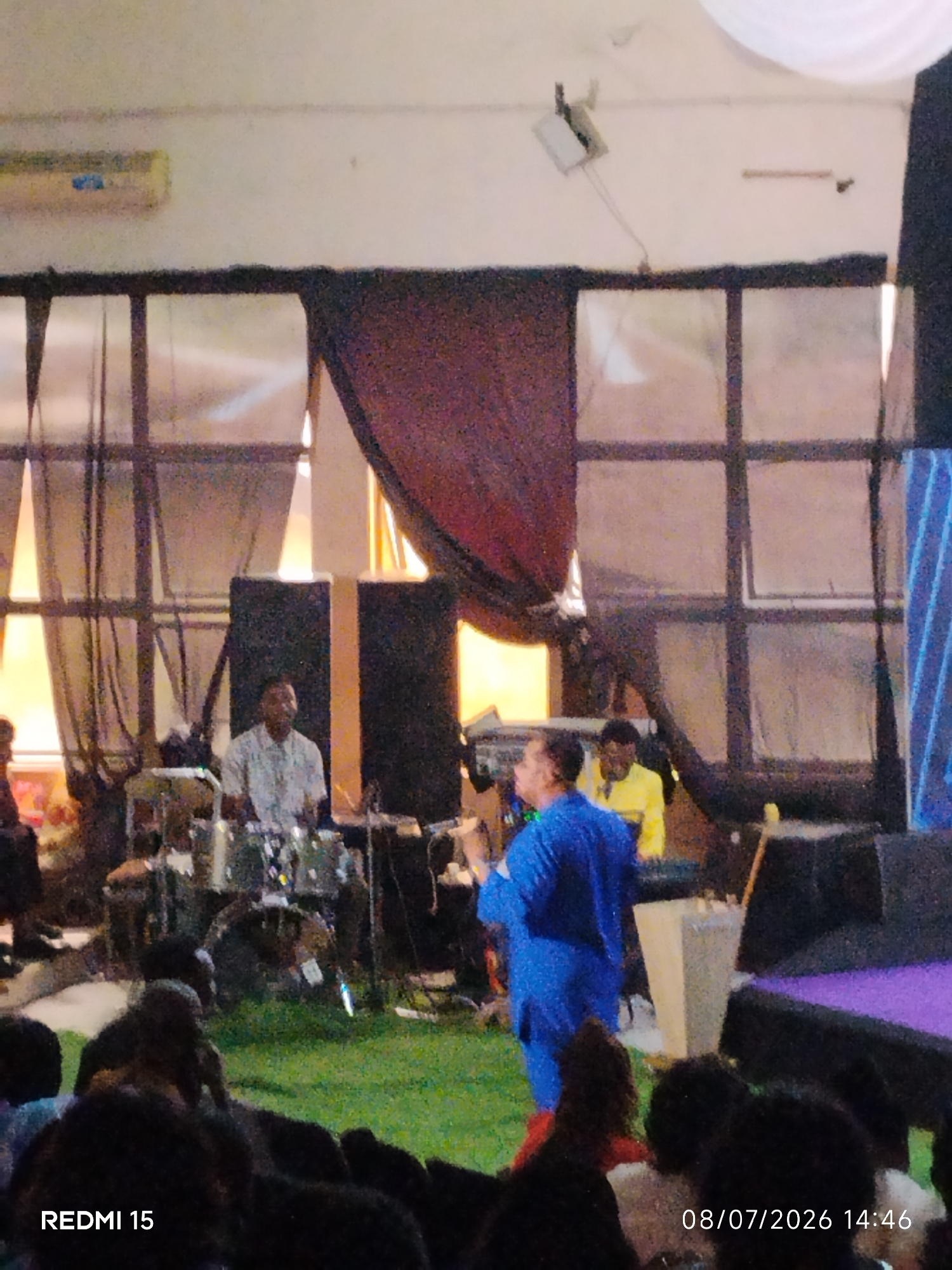 A group of men are gathered in what appears to be an indoor event space, possibly a church or community hall, in Aba, Nigeria. The scene is illuminated by artificial lighting. In the foreground, an audience is partially visible, seated on a grassy surface. In the mid-ground, a drummer is playing, seated behind a drum set. To the right of the drummer, a man wearing a blue outfit is speaking or singing into a microphone. Behind him, two other men are standing, one wearing a yellow shirt. The background features large windows with dark frames, covered by curtains. The overall atmosphere suggests a live performance or religious service. The timestamp "08/07/2026 14:46" is present in the bottom right corner of the image.