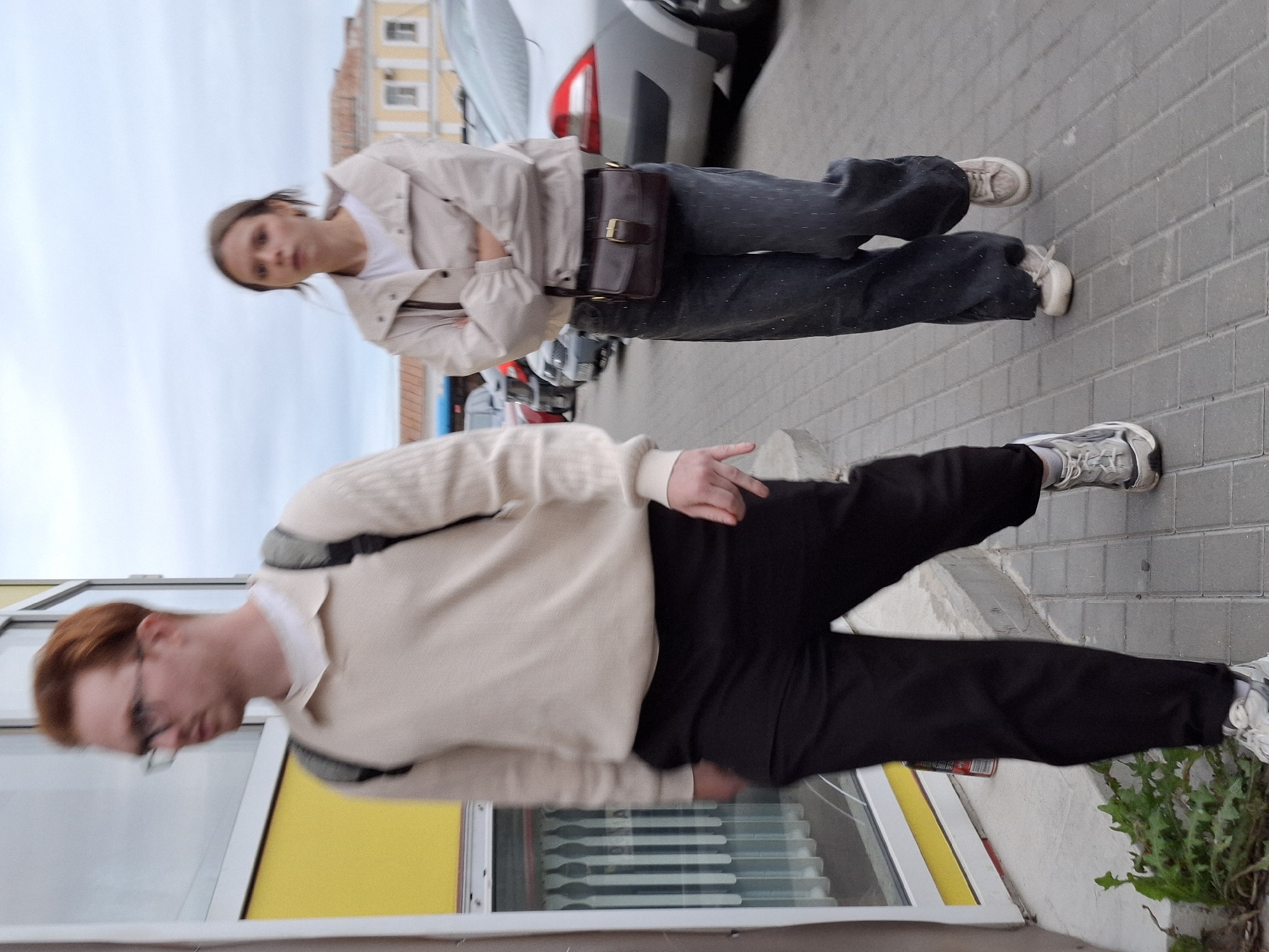 A man with red hair and glasses, wearing a light-colored sweater, black pants, and a backpack, walks on a sidewalk. A woman with brown hair, wearing a light-colored jacket, dark pants, and a brown crossbody bag, walks behind him. The background shows parked cars and buildings under a cloudy sky.