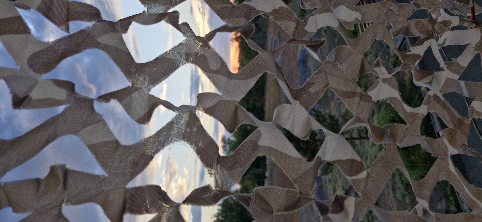 The image is a close-up shot of a camouflage net against a backdrop of a sunset sky and a rural landscape. The net has a pattern of tan and beige irregular shapes, with frayed edges suggesting it is made of fabric. Through the openings in the net, a cloudy sky with hues of orange and pink from the setting sun is visible. Below the sky, a line of trees and green foliage can be seen, with a patch of bare ground and what appears to be a blue tarp or structure in the middle distance. The foreground is dominated by the camouflage net, which obscures much of the view of the environment. The image provides no specific information about the city of Usharal, Kazakhstan, or any people or actions within the scene.