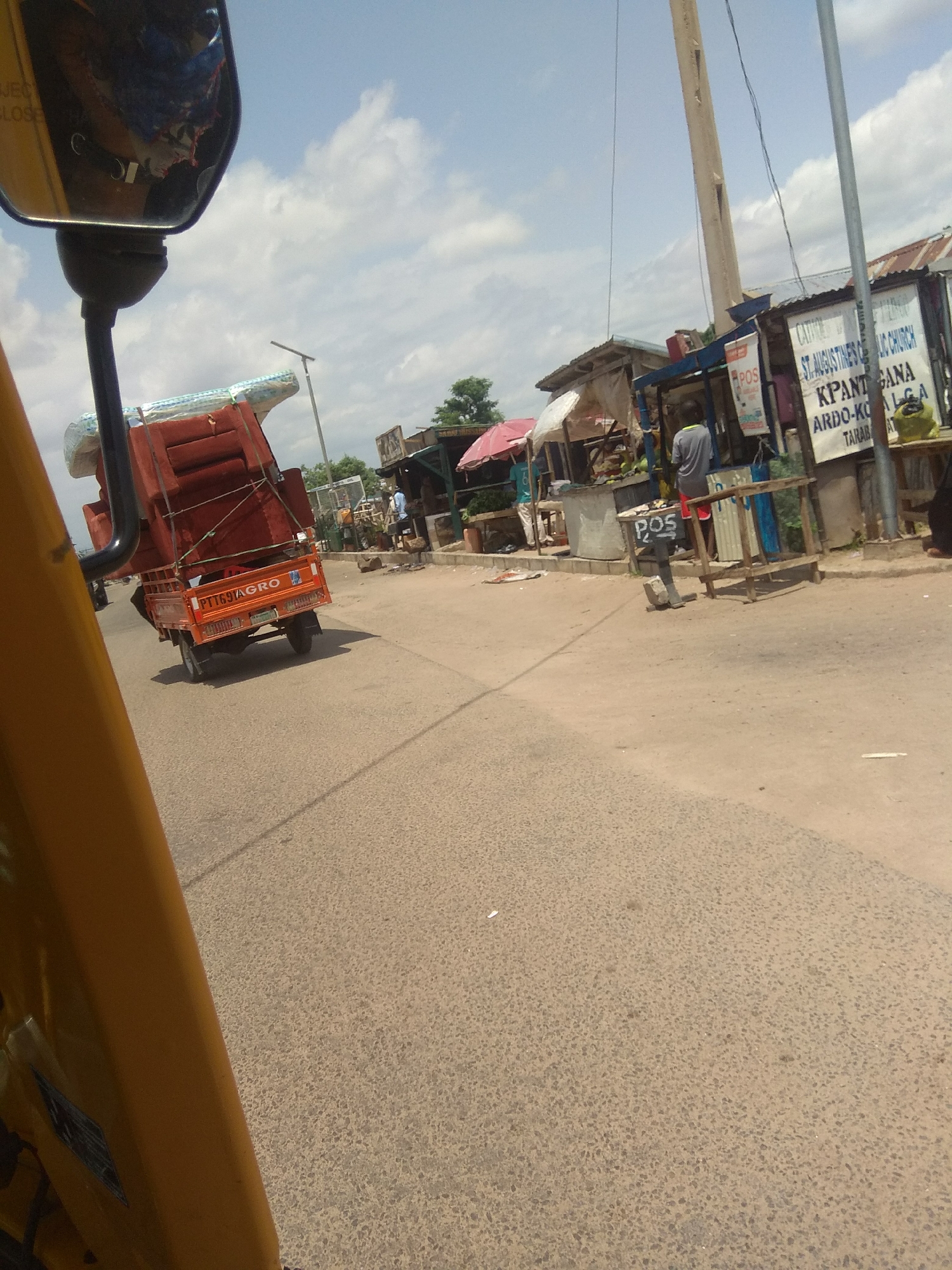 This is a media file showing a street scene in Jalingo, Nigeria. The foreground is dominated by the yellow-orange structure of a vehicle, possibly an auto-rickshaw or tricycle, from which the photo is taken. The reflection in the side mirror shows a blue and red object, partially obscured.
The main subject in the middle ground is an orange three-wheeled vehicle carrying a large load of what appears to be furniture, possibly chairs or sofas, wrapped in plastic and secured with ropes. The vehicle has a license plate that reads "PTT691AGRO".
To the right of the vehicle, there are several stalls or small shops lining the street. One stall has a sign with text including "ST AUGUSTINES CELIC CHURCH", "KPANT GANA", and "ARDO-KO ALGA". Another stall has a "POS" sign. A person in a grey shirt and red shorts is standing near one of the stalls. Another person in a blue shirt and white pants is visible in the distance.
The scene appears to be set during the day, under a partly cloudy sky. The road is paved with asphalt. The overall impression is of a busy market or roadside commercial area. The visible text provides specific location details related to a church and possibly a local government area.