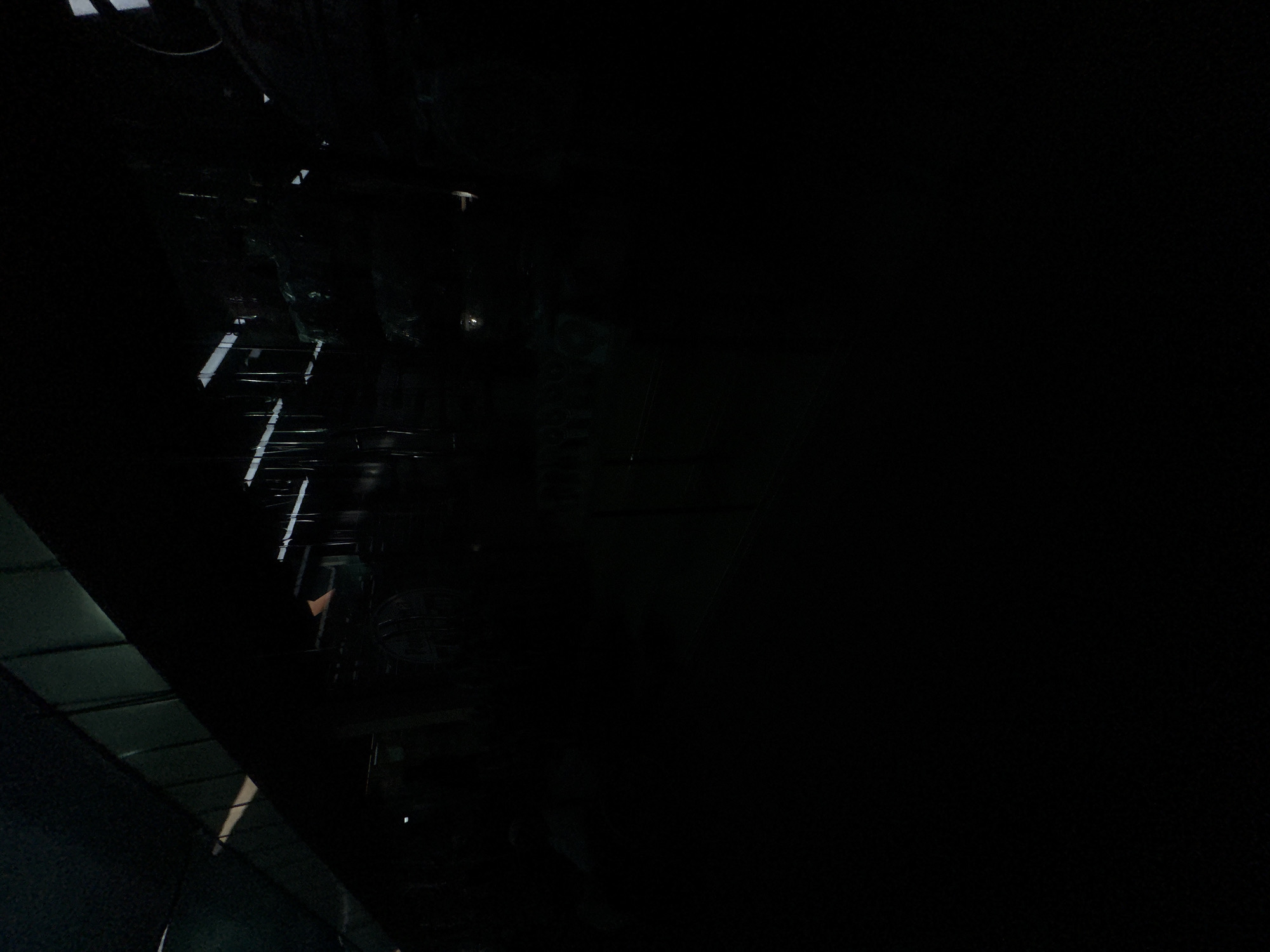 The image shows a dimly lit interior, possibly a warehouse or factory. The ceiling is visible in the upper left, with metal beams and panels. In the mid-ground, there are structures that appear to be machinery or shelving, and some circular objects that could be barrels or tanks. Lights are present on the ceiling and some are also visible in the background. The lower portion of the image is entirely black, indicating that this area is not illuminated or is out of frame. The overall impression is one of an industrial or storage space. There are no people visible in the image. The crops provided offer closer views of specific areas within the scene, revealing details such as the texture of materials and the presence of various industrial elements.