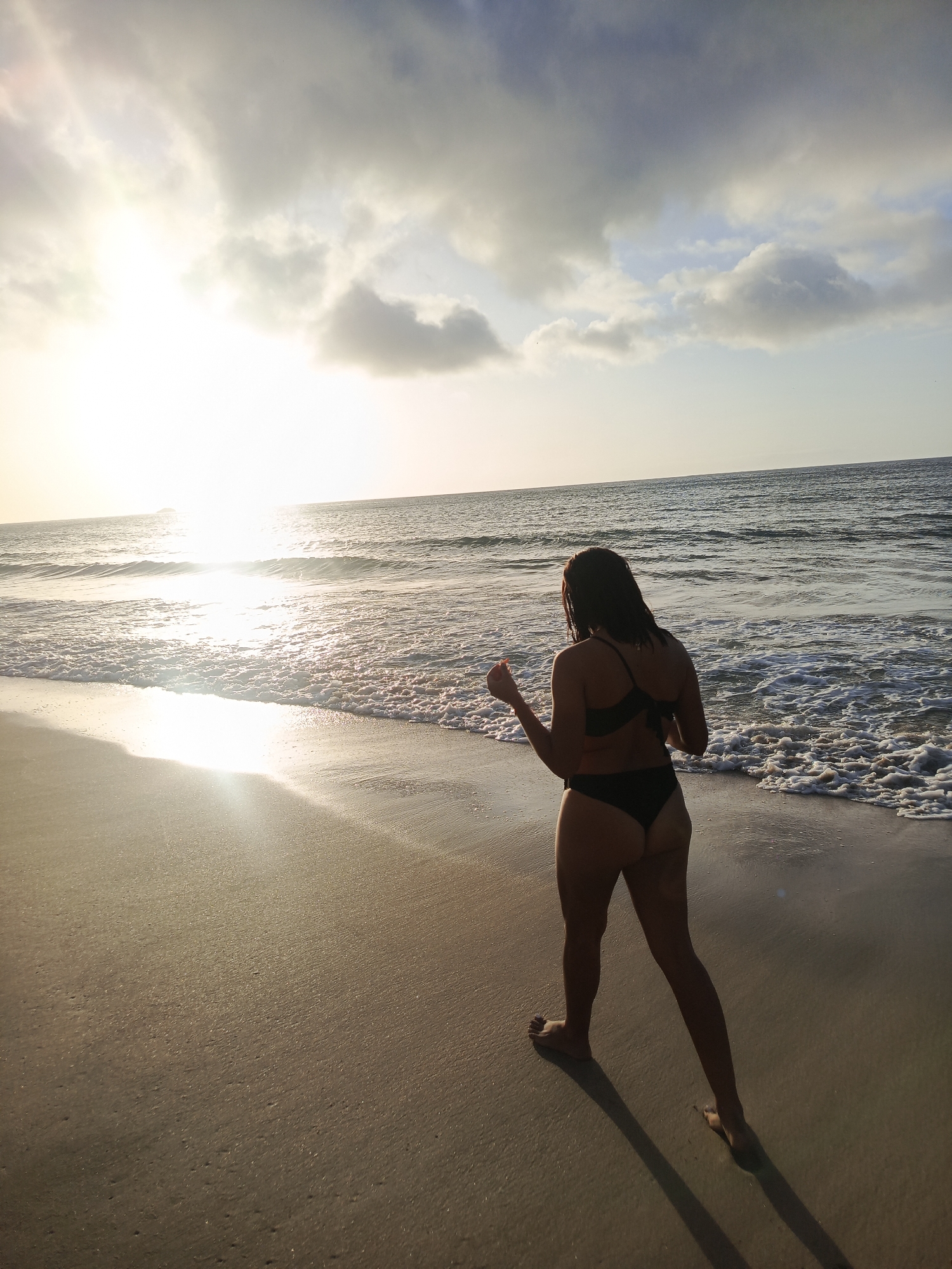 A person wearing a black bikini stands on a sandy beach, facing away from the camera and towards the ocean. The sun is setting, casting a bright glare on the water and creating long shadows on the sand. Gentle waves are breaking on the shore. The sky is partly cloudy with the sun peeking through. The location is the coast of Pedro González, Venezuela.