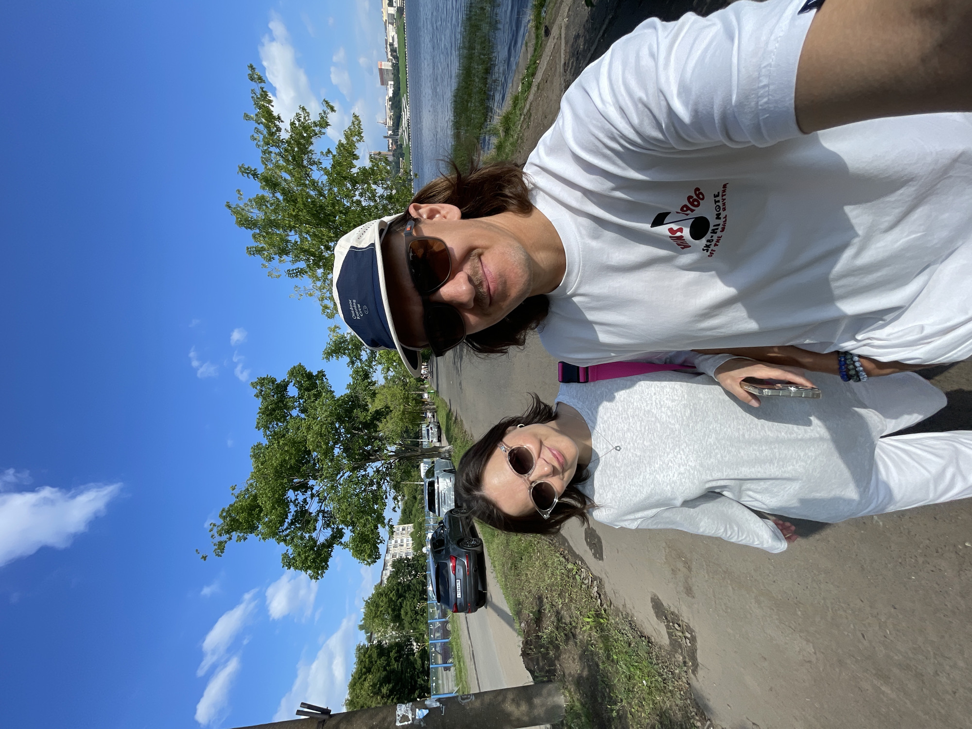 Two individuals, a man and a woman, are taking a selfie on a paved walkway next to a body of water. The man is in the foreground, wearing sunglasses, a white t-shirt with graphics, and a cap. The woman is to his left, wearing sunglasses and a light-colored outfit. Behind them, cars are parked on the side of a road, and trees line the path. In the distance, buildings and a cityscape are visible across the water. The sky is blue with scattered clouds. The scene appears to be a recreational area within the city of Izhevsk, Russia, possibly along the Izh River or the Izhevsk Reservoir.