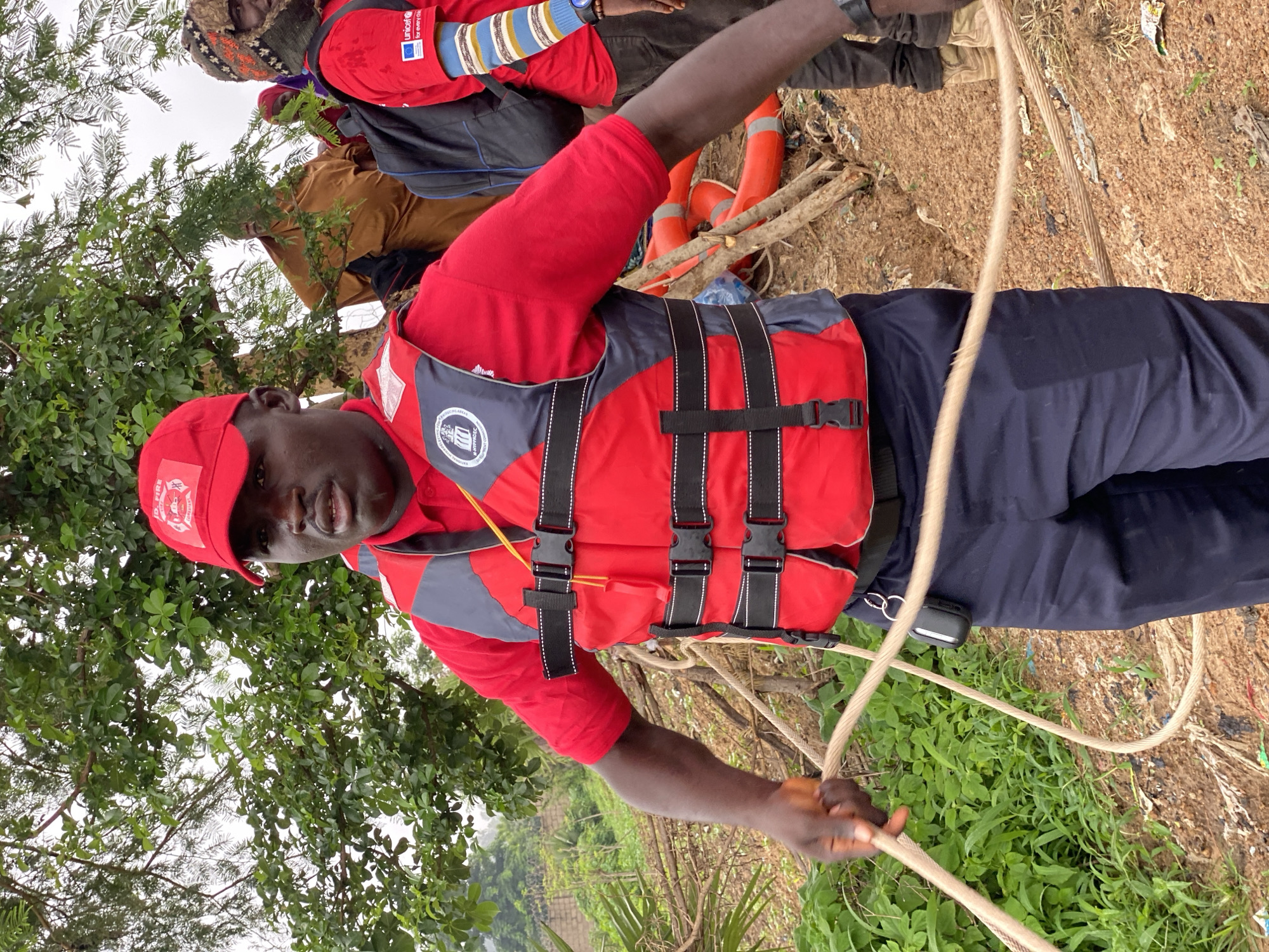 In Kawo, Nigeria, a man in a red shirt and life vest is pulling a rope. He is wearing a red cap with a badge that says "D. FIRE COMMAND". In the background, other people are visible, including one wearing a brown robe and another with a UNICEF shirt and colorful arm bands. The ground is covered in dirt, debris, and green foliage, with trees and branches in the background. There are also orange life preservers on the ground.