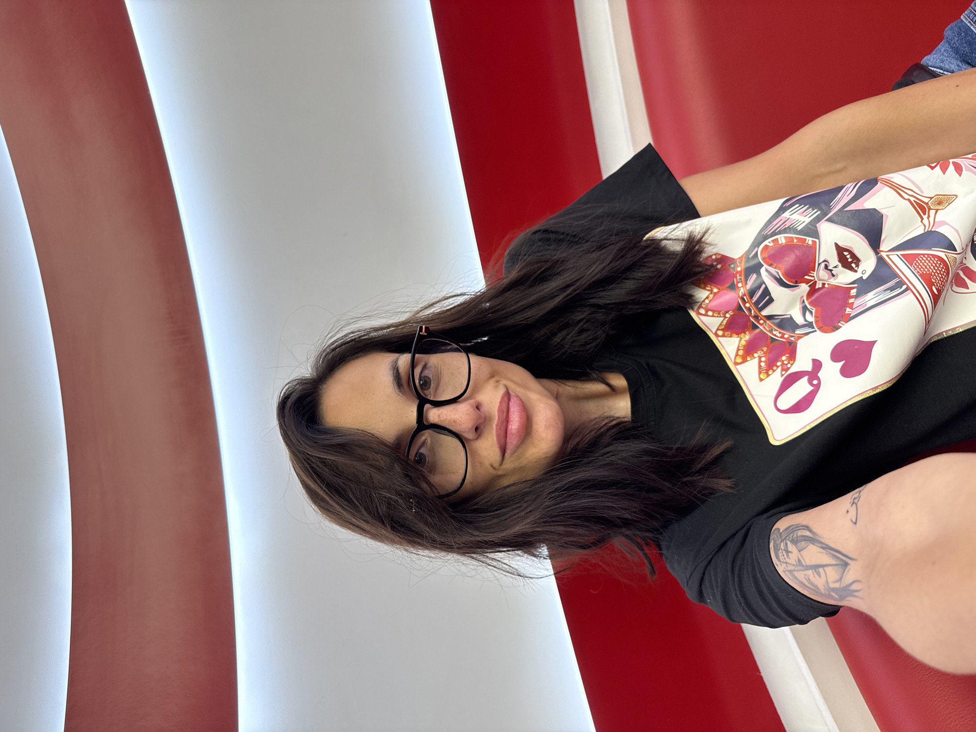 A woman with long brown hair and glasses is seated on a red and white striped structure, possibly a bench or wall, with white stripes illuminated. She's wearing a black t-shirt with a graphic print of the Queen of Hearts playing card and has a tattoo on her inner left arm. The location is in Pleven, Bulgaria.