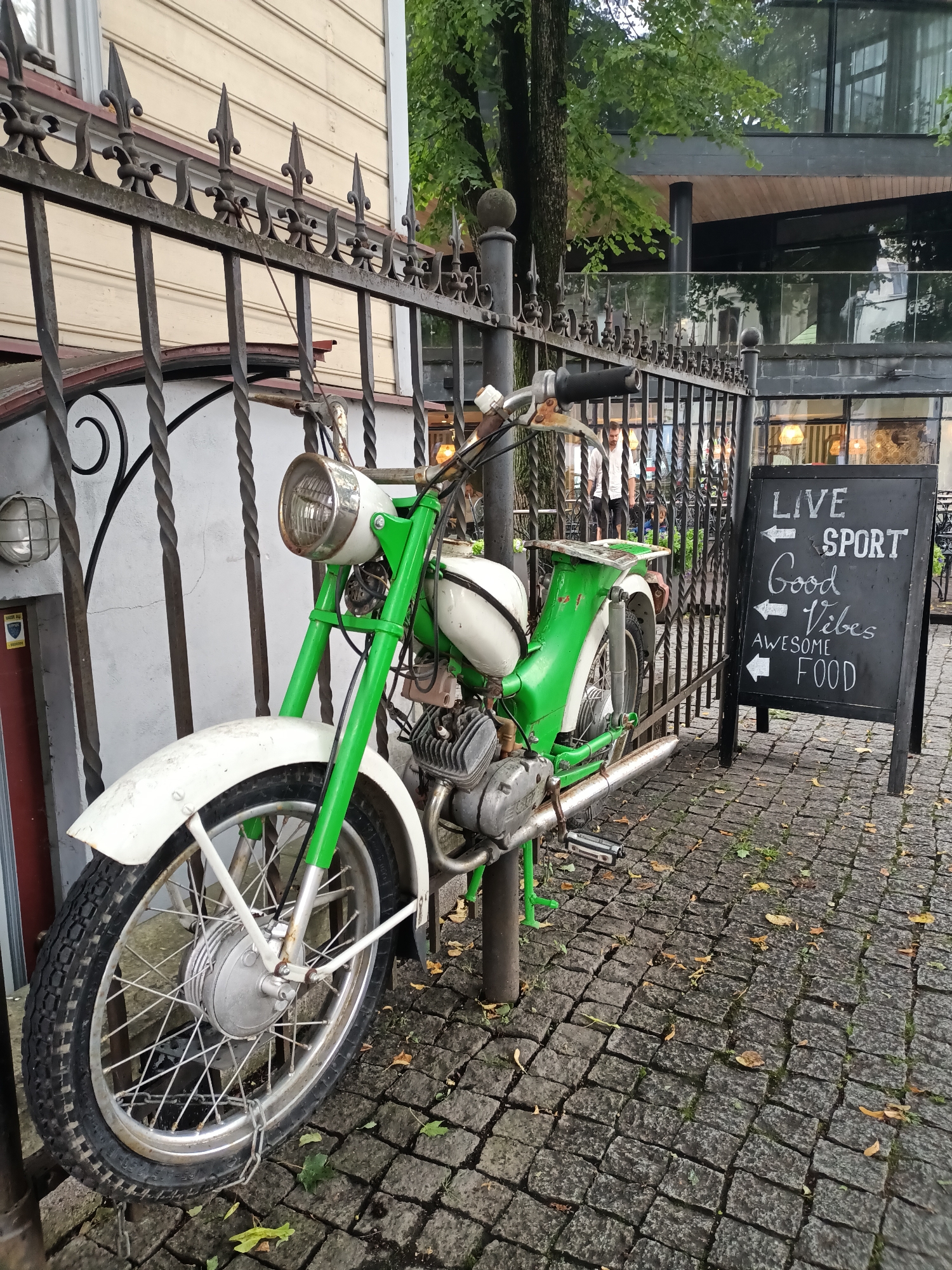 A vintage green and white moped is parked next to a black chalkboard sign on a cobblestone street in Tallinn, Estonia. The moped is leaning on its kickstand, secured by a chain wrapped around its front wheel and a metal pole. The chalkboard sign has white lettering that reads "LIVE SPORT Good Vibes AWESOME FOOD" with arrows pointing left next to each line. Behind the moped and fence, a modern building with glass windows and balconies is visible, along with trees and foliage. The scene appears to be outdoors, possibly in a public area or outside a cafe or restaurant.