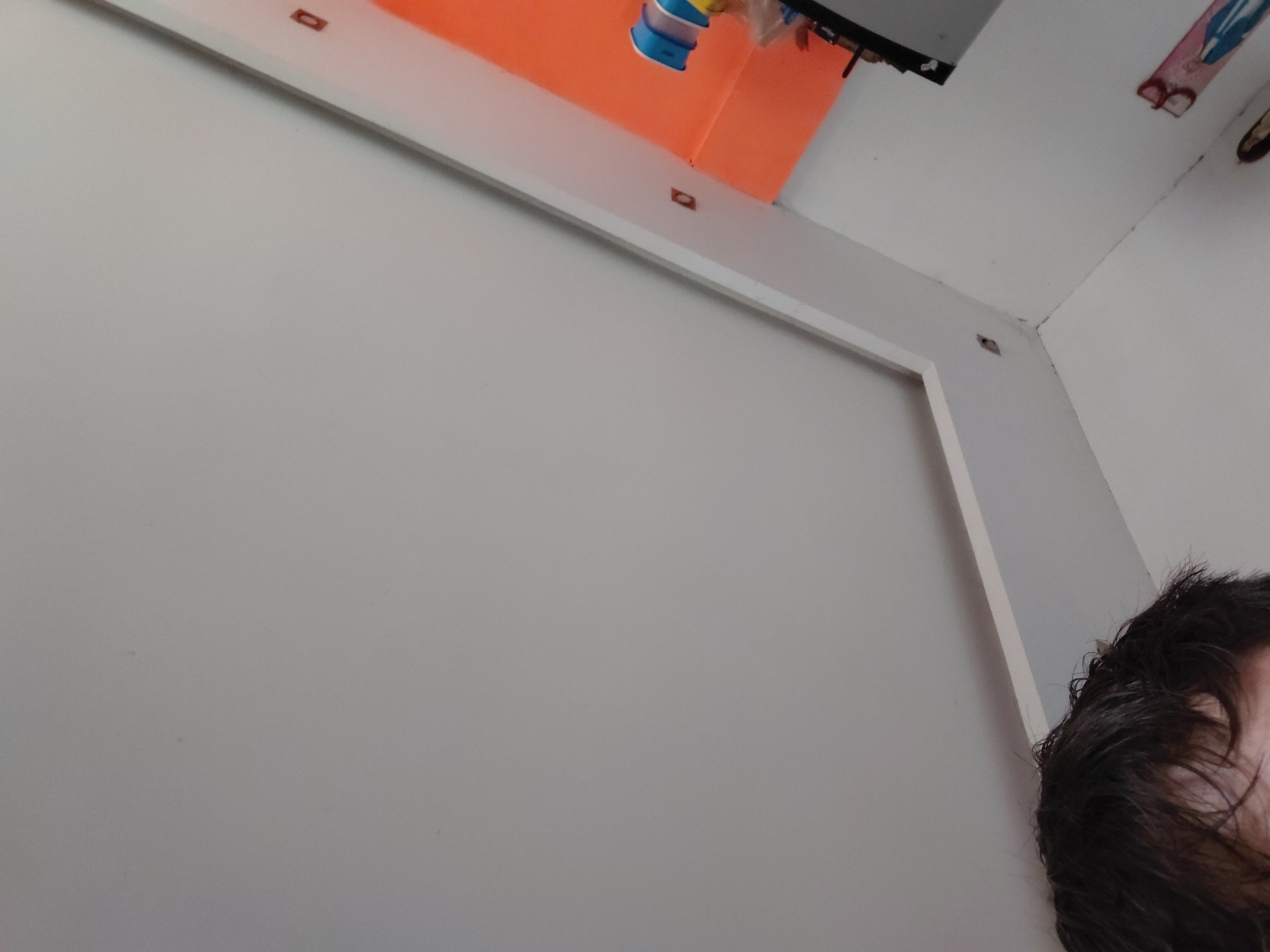 The image captures an indoor scene, likely a room or a large piece of furniture, from a low-angle perspective. The dominant feature is a large, light grey or off-white vertical surface, possibly a wall or a closet door, filling most of the left and central frame.
Above this surface, a white horizontal beam or frame extends across the image. Behind this beam, an orange-colored surface is visible, possibly another wall or a large object. In the upper-central area, a blue and yellow cylindrical object, resembling a toy or dispenser, is partially visible, seemingly attached or placed near the orange surface. Small, dark, rectangular-shaped objects, possibly fasteners or decorative elements, are intermittently spaced along the white beam.
The top right corner shows a white ceiling and an adjoining wall with fragmented views of decorative items, including one with pink and blue patterns and another dark, circular element. In the bottom right corner, a portion of a person's head with dark, somewhat disheveled hair is visible, suggesting a close proximity to the camera or a first-person perspective. No facial features or discernible emotions are apparent.
There is no specific activity taking place, and no legible text, numbers, or branding are visible within the frame. The lighting is even, suggesting an indoor setting without strong natural light indicators for time of day.