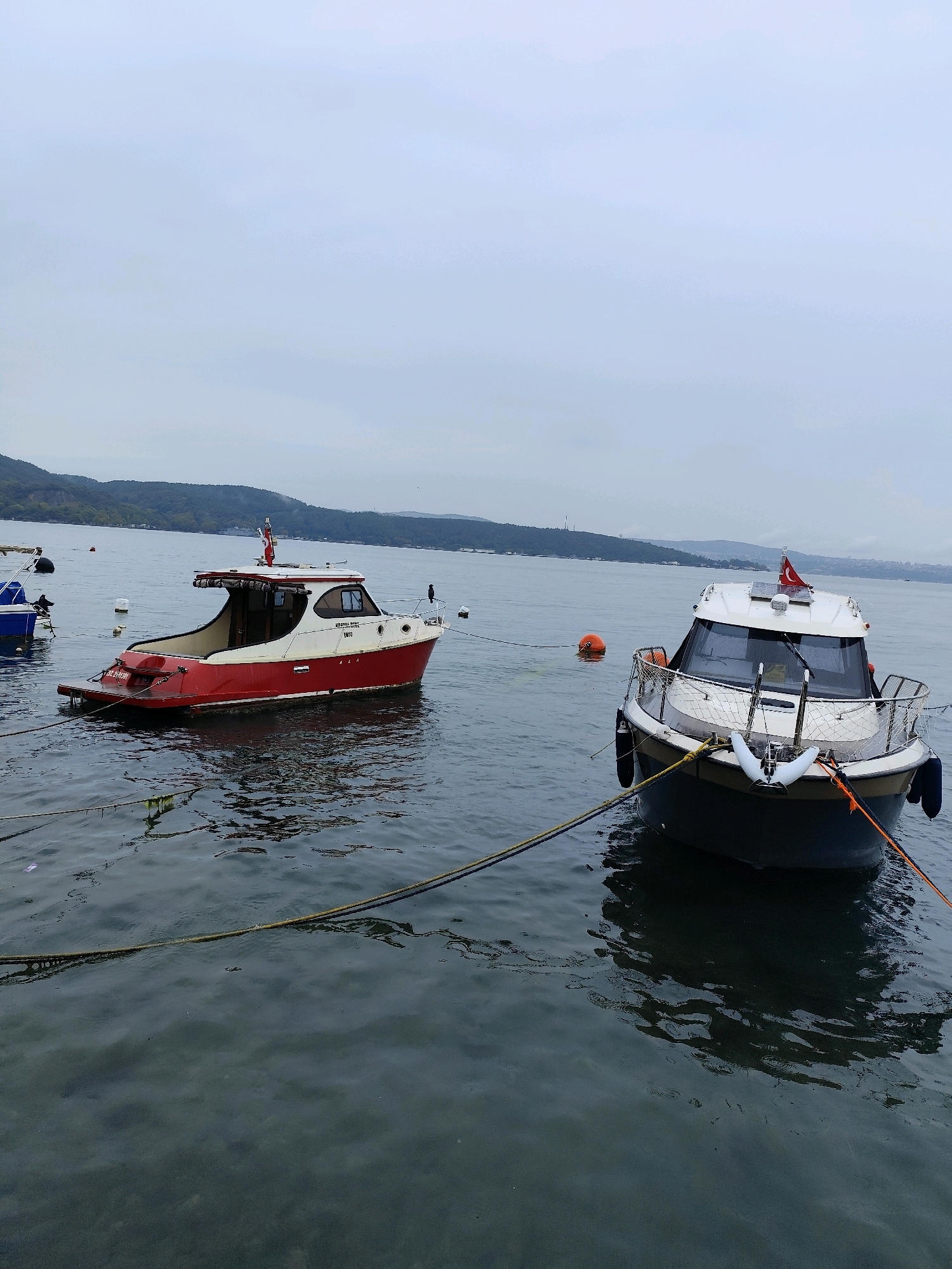 The image shows a cloudy day at a waterfront location in Sarıyer, Turkey, likely the Bosphorus strait. Two boats are moored in the water. The boat on the left is red and white with a covered cabin and an open stern deck. The boat on the right is white and grey with a more modern design, featuring a cabin with large windows and a Turkish flag flying from the roof. Both boats are secured with ropes, and buoys are visible in the water. In the background, a hilly, tree-covered shoreline can be seen, typical of the Sarıyer district. The water appears calm with some ripples. The overall scene depicts a serene, possibly overcast, maritime environment.